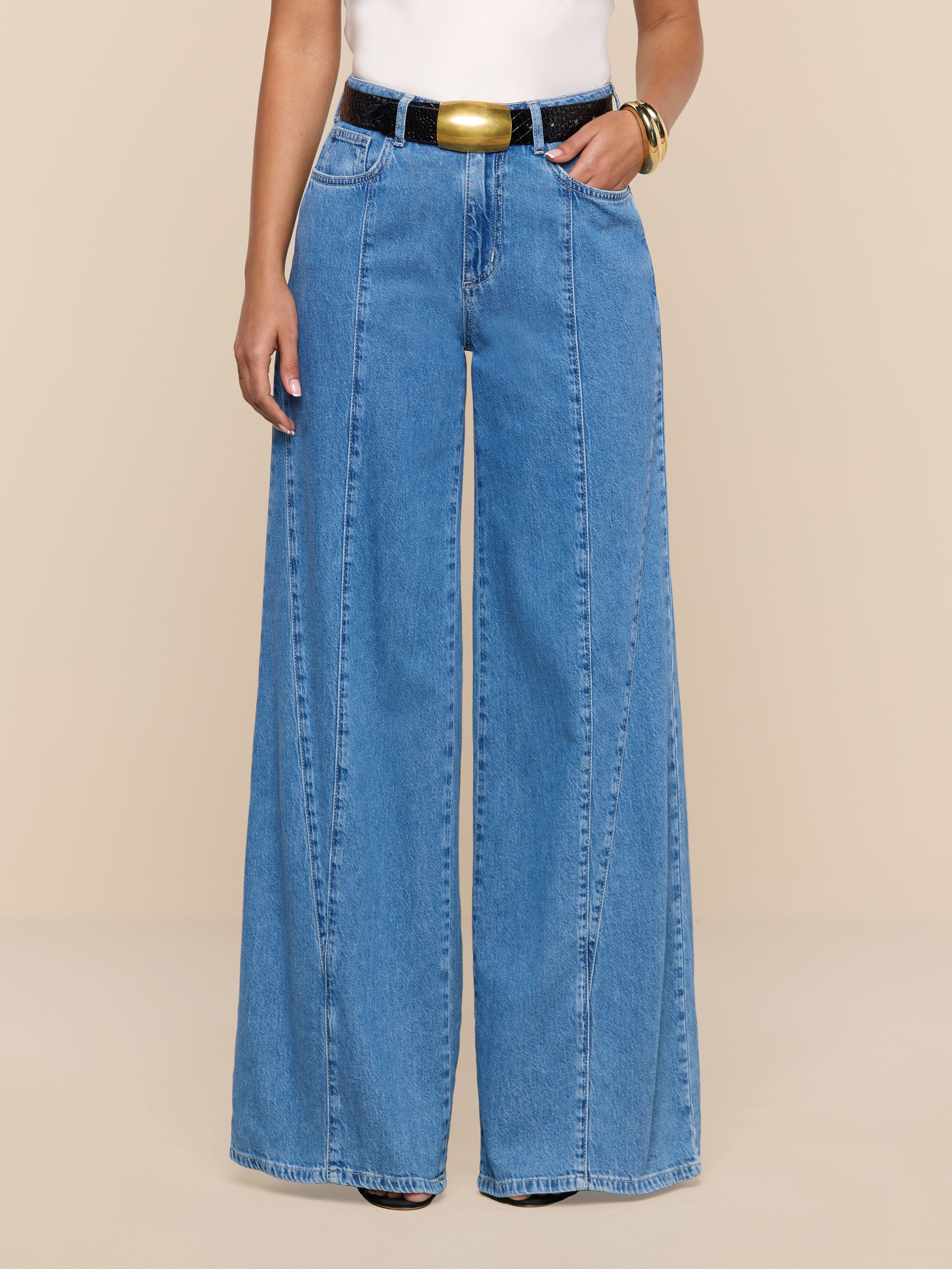 A person wearing the Niles Wide-Leg Jean, featuring a high-rise, wide-leg silhouette in lightweight blue denim, styled with a black belt and white top, stands against a plain white background.