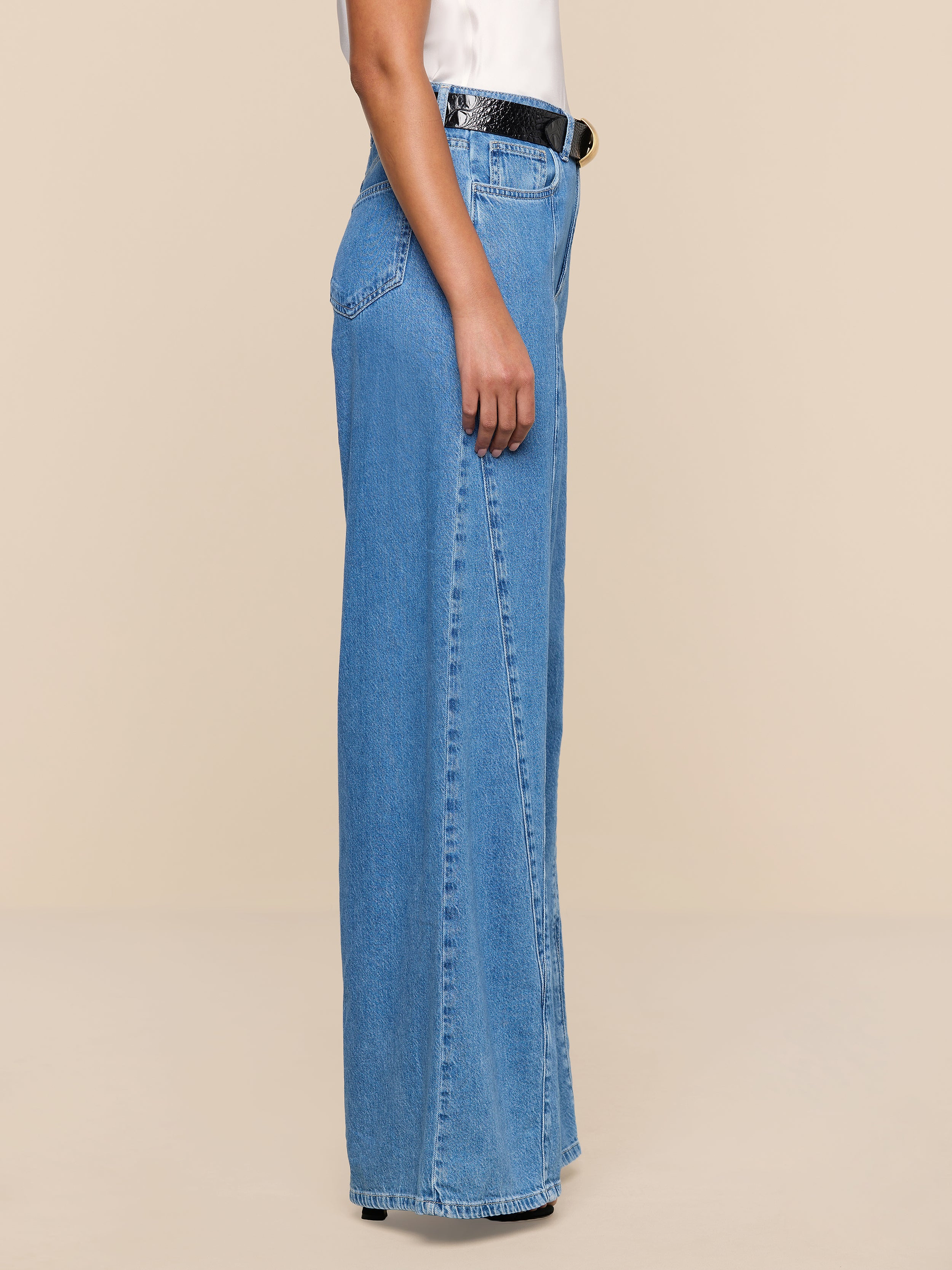 Sure Here’s the revised sentence using your product name: A person stands sideways against a plain white background, wearing a white top tucked into high rise Niles Wide-Leg Jeans made from lightweight blue denim, styled with a black belt.