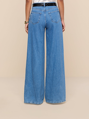A person is shown from the back wearing the Niles Wide-Leg Jean—high-rise, all-cotton denim with wide legs, two back pockets, and frayed hems—posing against a plain beige background for a relaxed look.