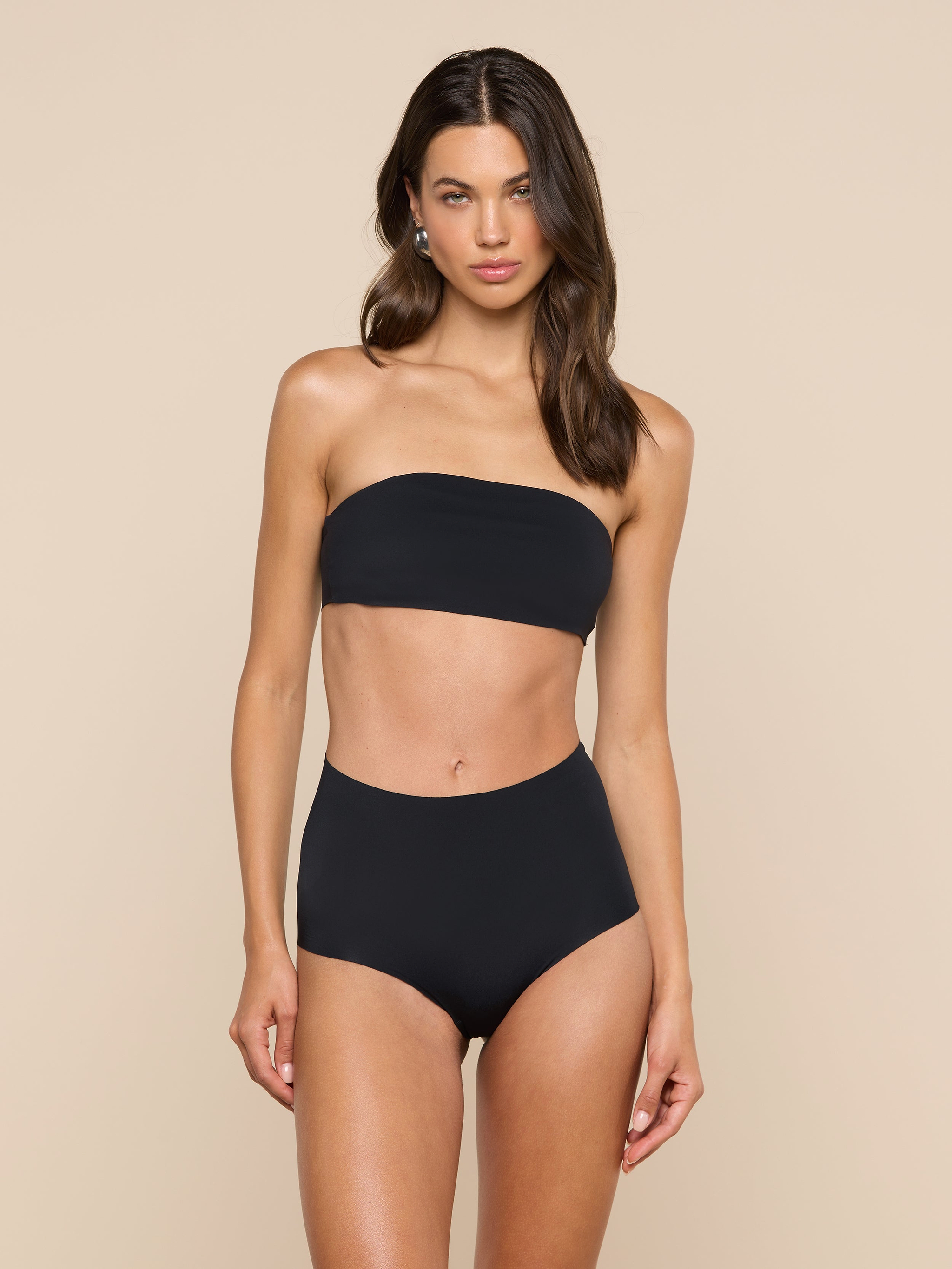 A woman stands and poses in the Velle Knit Panty paired with a black strapless bandeau bikini top against a plain, light background.