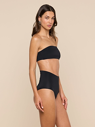 A woman stands and poses against a plain light background wearing the Velle Knit Panty in black, styled as part of a strapless bandeau bikini set with high-waisted bottoms.