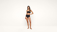 A woman stands barefoot in a black strapless bikini against a plain white background, looking at the camera with one hand resting on her hip.