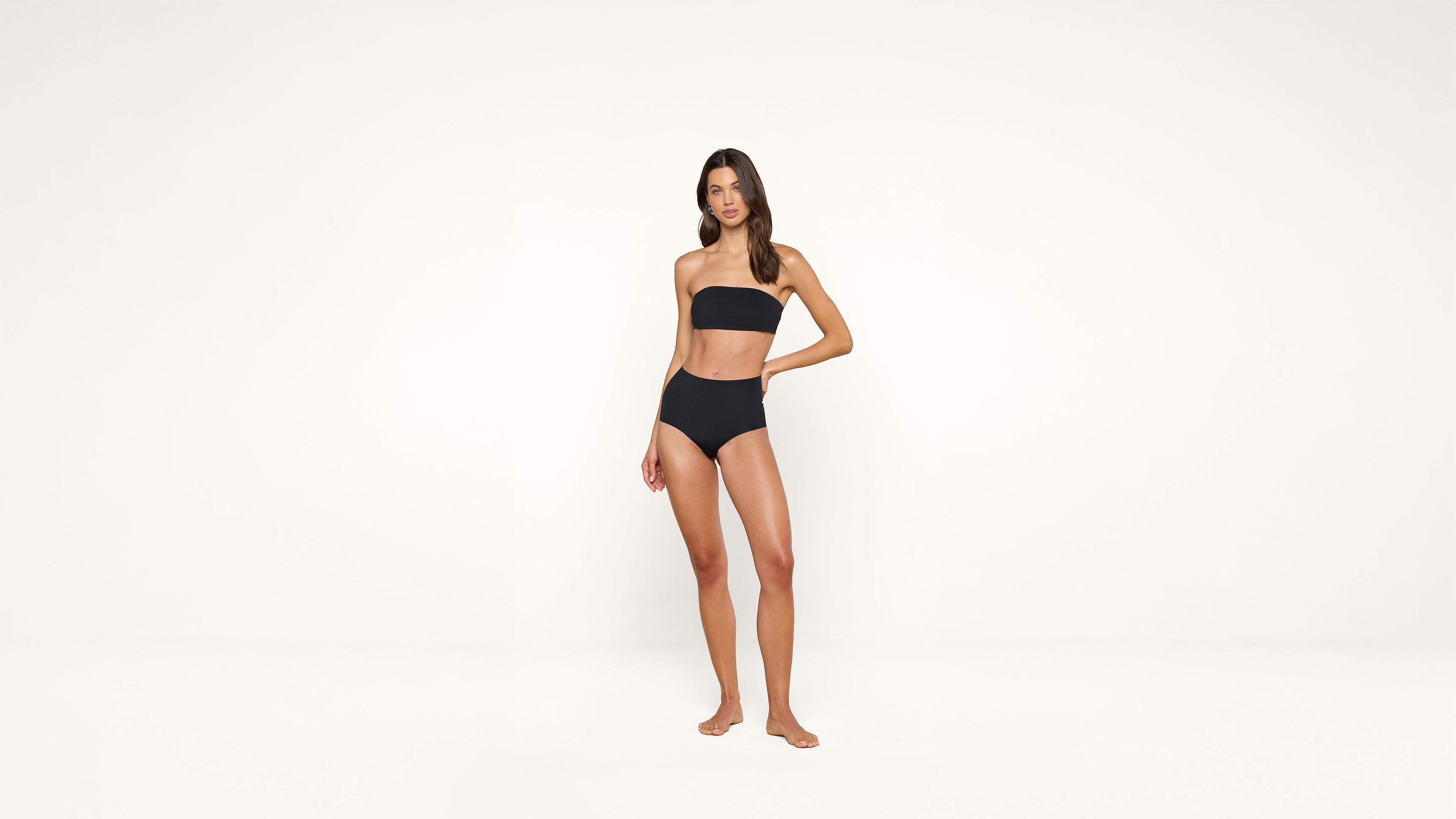 A woman stands barefoot in a black strapless bikini against a plain white background, looking at the camera with one hand resting on her hip.