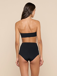 A woman with long brown hair, seen from the back, wears a black strapless bandeau top and the Velle Knit Panty against a plain light background.