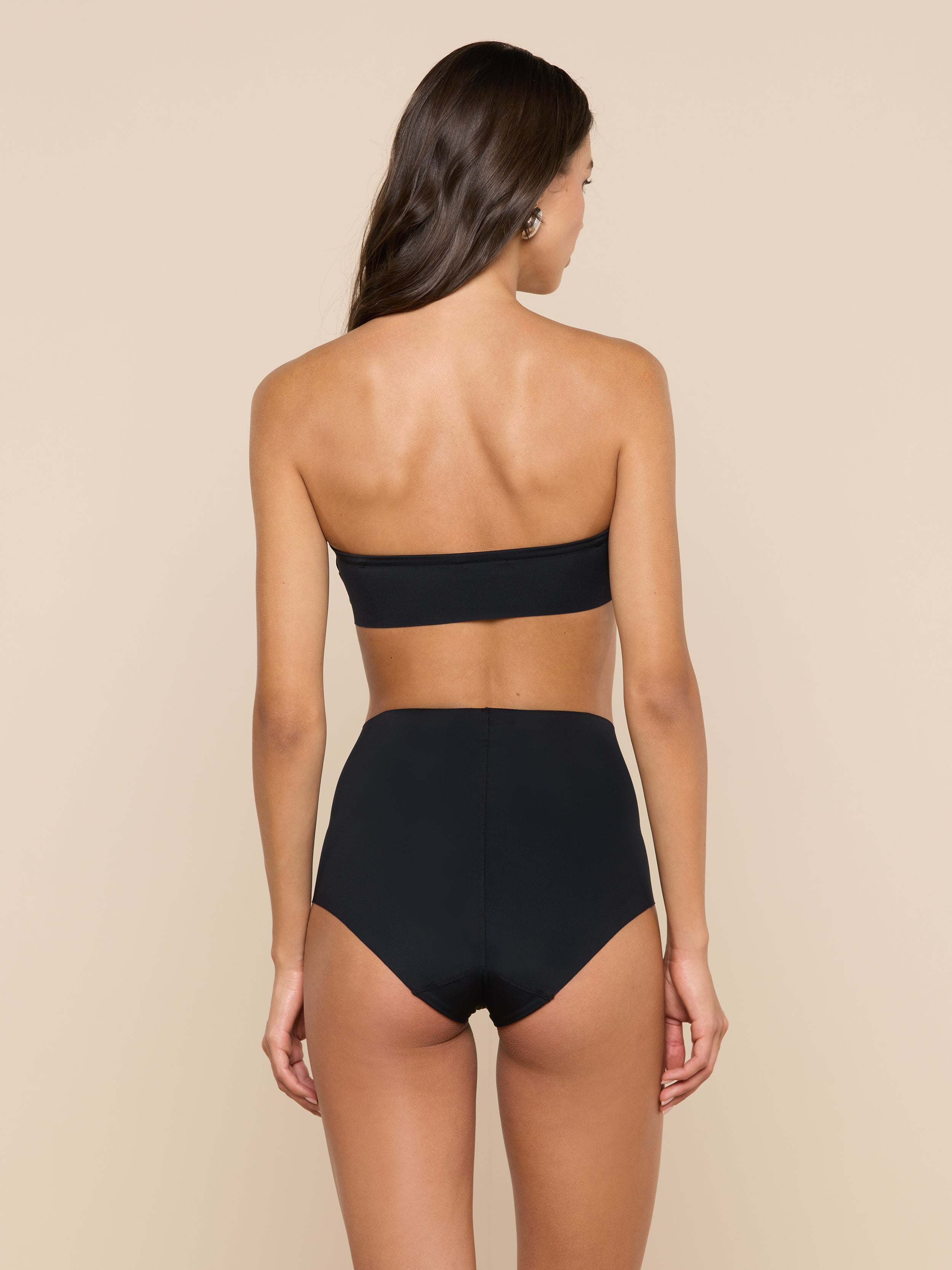 A woman with long brown hair, seen from the back, wears a black strapless bandeau top and the Velle Knit Panty against a plain light background.