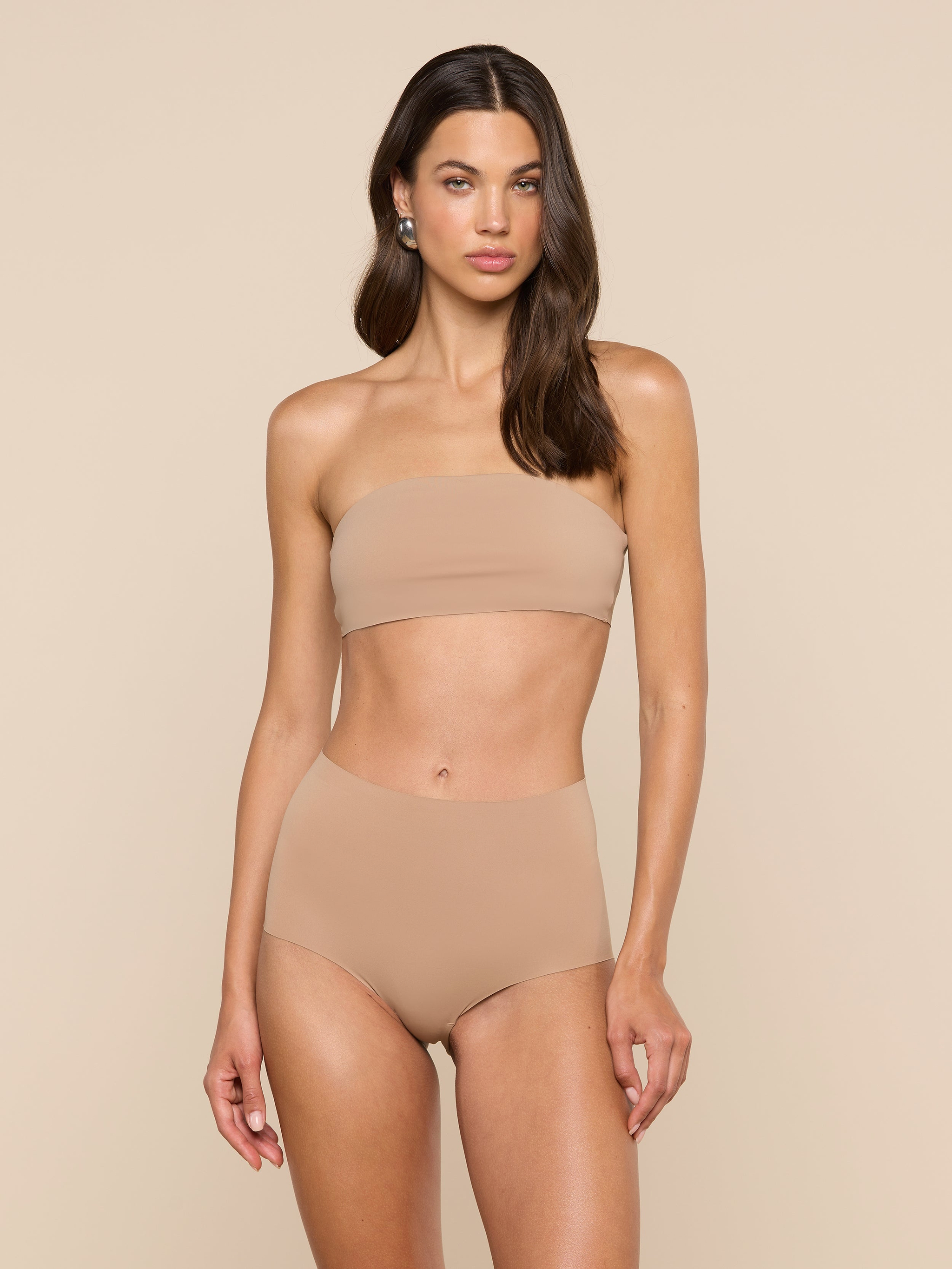 A woman with long, wavy hair and a neutral expression stands against a plain background wearing the Velle Knit Panty paired with a strapless nude bandeau bikini top.