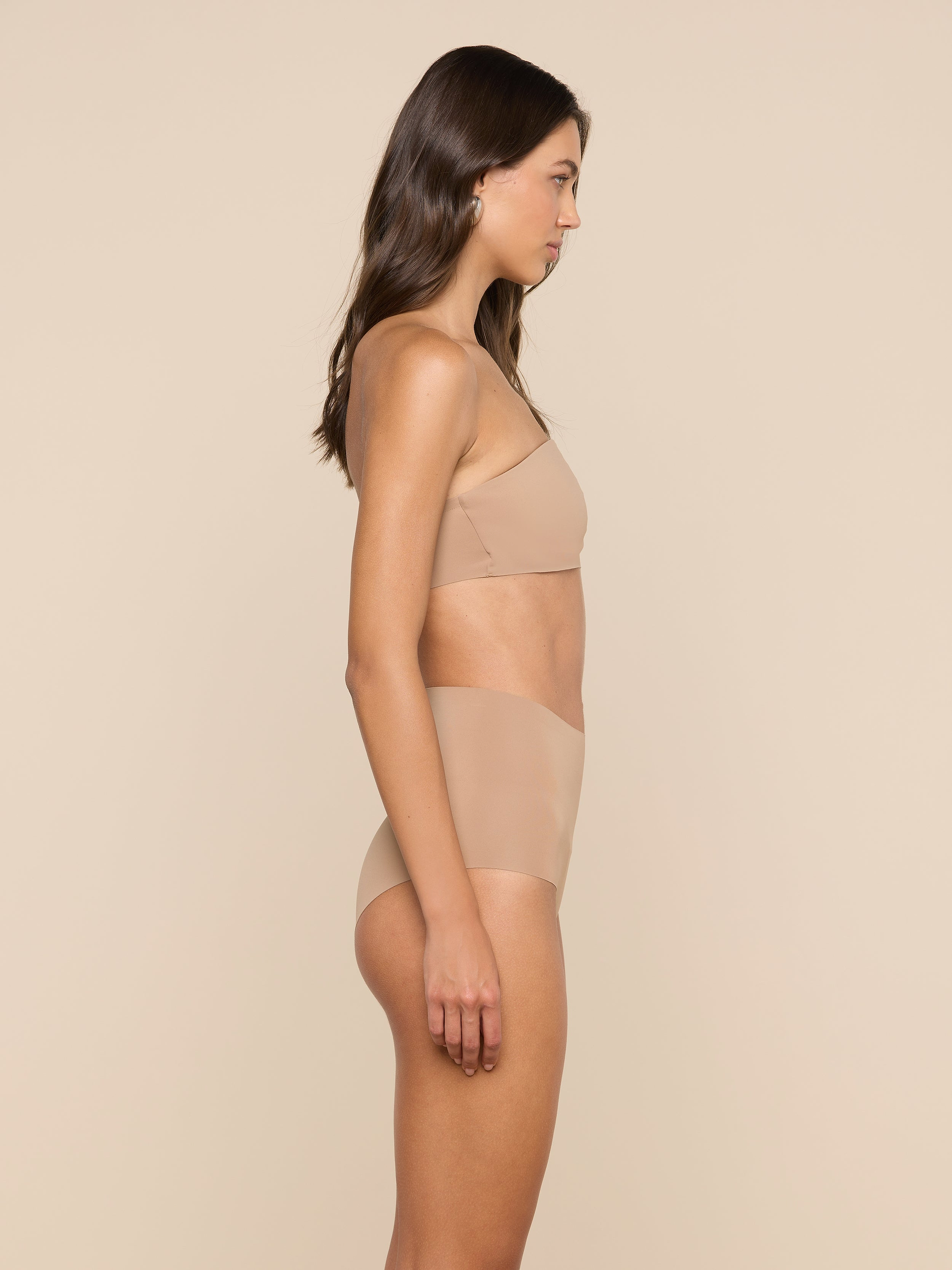 A woman stands in profile against a plain background, wearing a matching nude-colored L’AGENCE Velle Knit Panty and undergarment set.
