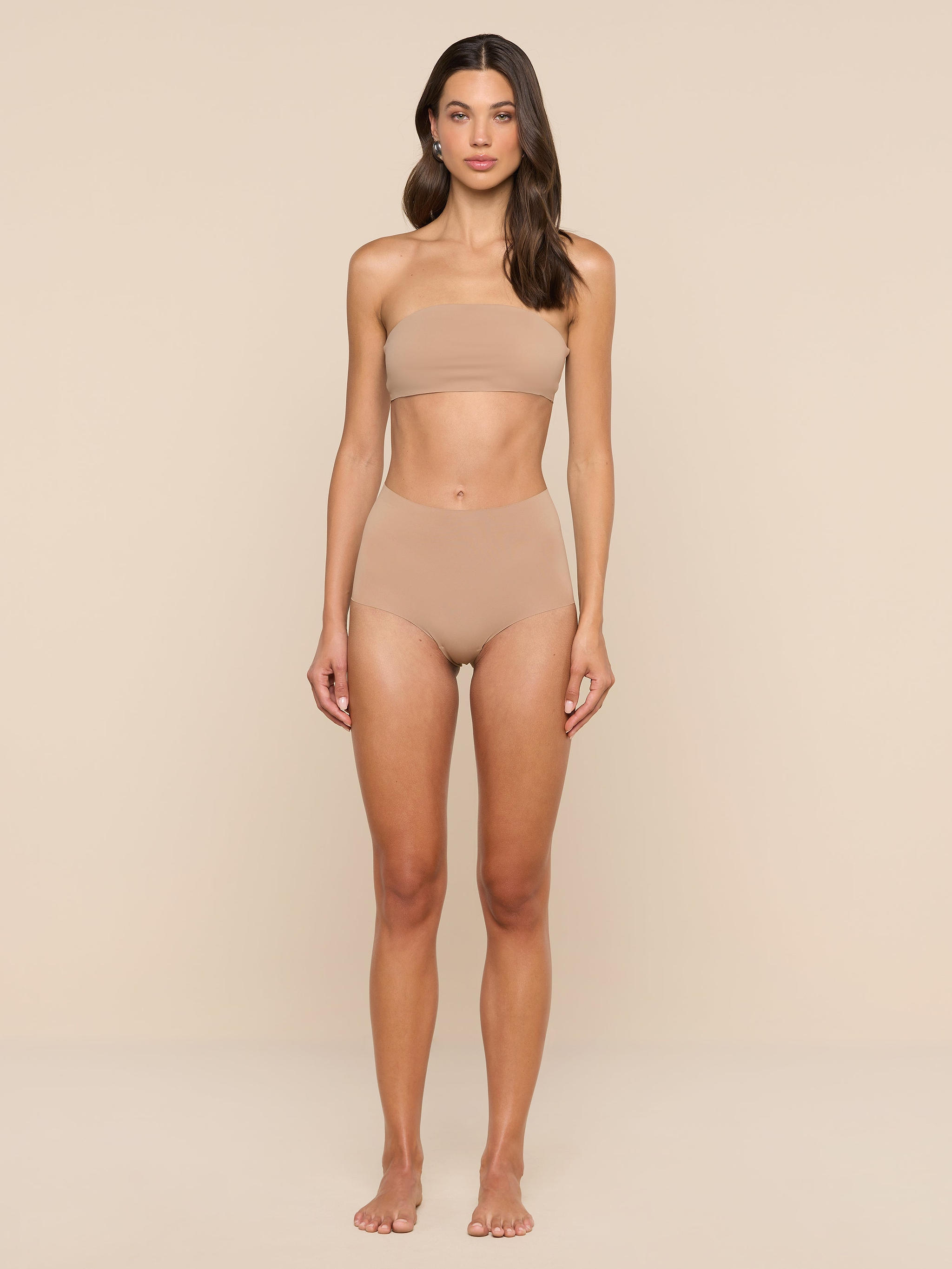 A woman stands facing forward, wearing a nude-colored bandeau top and the Velle Knit Panty, against a plain white background.
