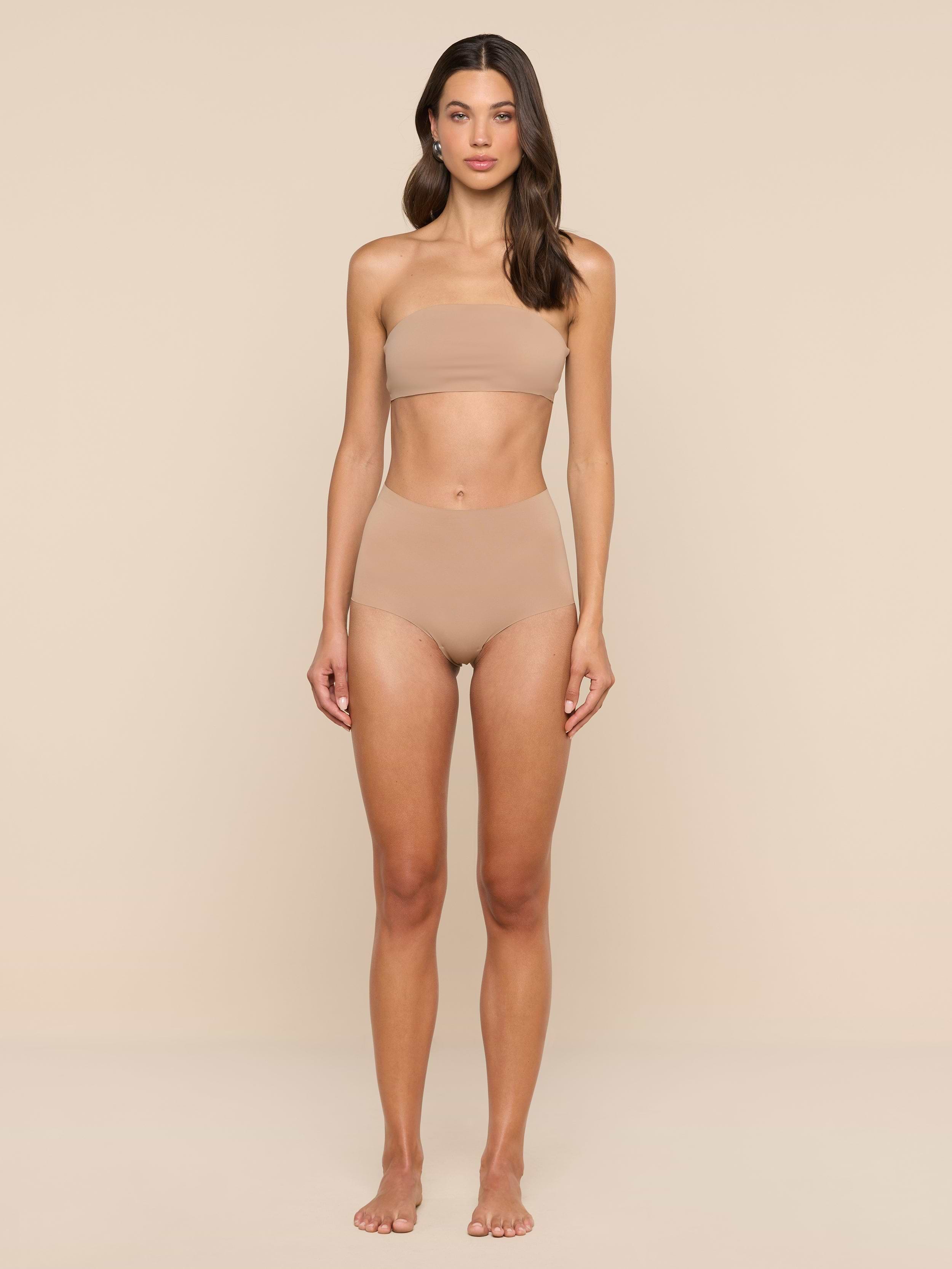 A woman stands facing forward, wearing a nude-colored bandeau top and the Velle Knit Panty, against a plain white background.