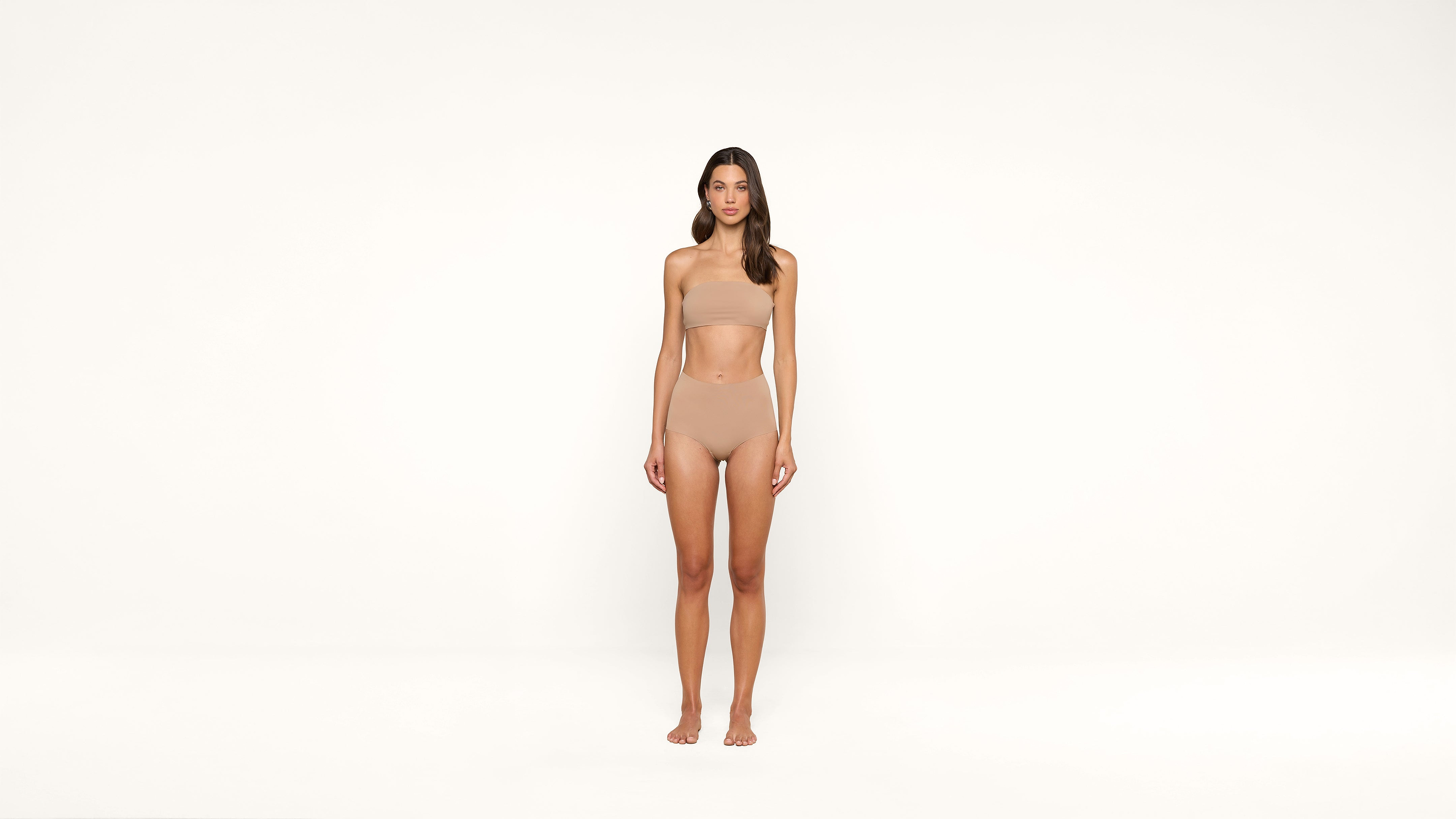 A woman stands barefoot against a plain white background, wearing seamless nude-colored underwear and a bandeau top. She faces forward with a neutral expression and her arms relaxed at her sides.