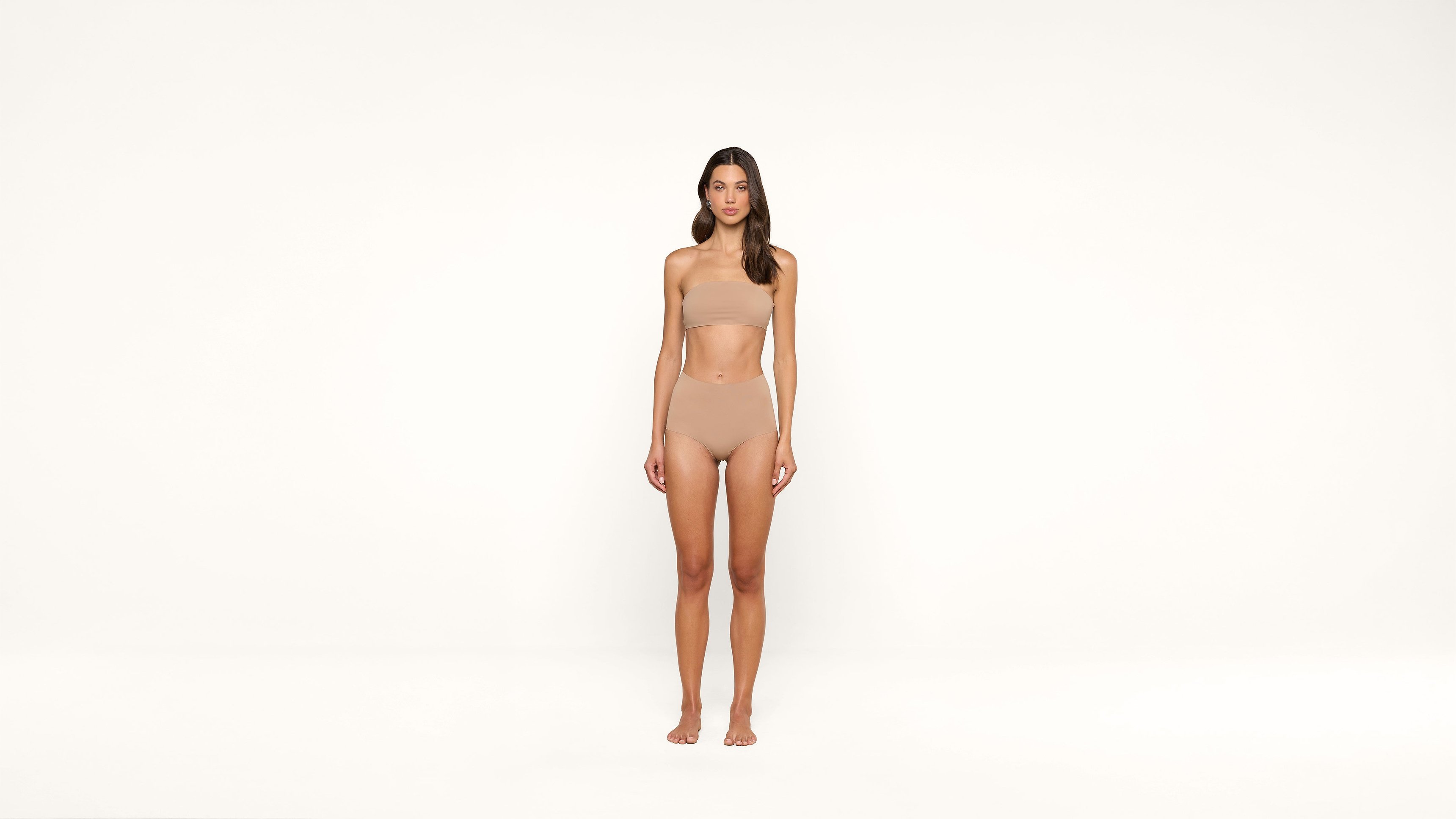 A woman stands barefoot against a plain white background, wearing seamless nude-colored underwear and a bandeau top. She faces forward with a neutral expression and her arms relaxed at her sides.