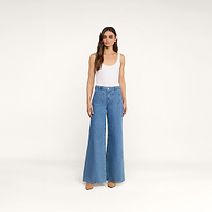 Une femme aux longs cheveux châtains se tient debout devant un fond beige uni, vêtue dun débardeur blanc sans manches et dun jean Delcan à jambes larges, à taille haute et doté de poches plaquées sur le devant, quelle associe à des chaussures couleur beige clair.