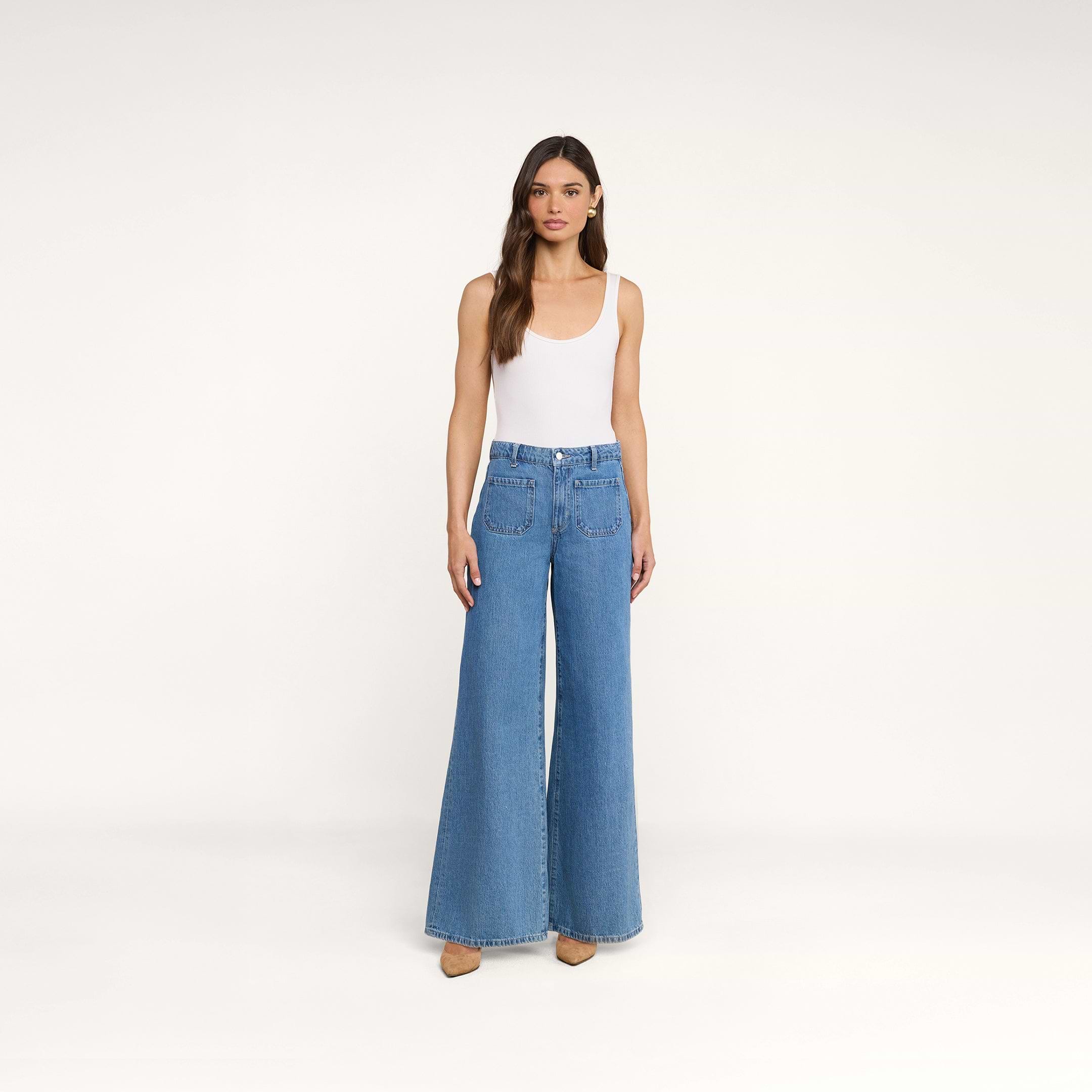 Une femme aux longs cheveux châtains se tient debout devant un fond beige uni, vêtue dun débardeur blanc sans manches et dun jean Delcan à jambes larges, à taille haute et doté de poches plaquées sur le devant, quelle associe à des chaussures couleur beige clair.