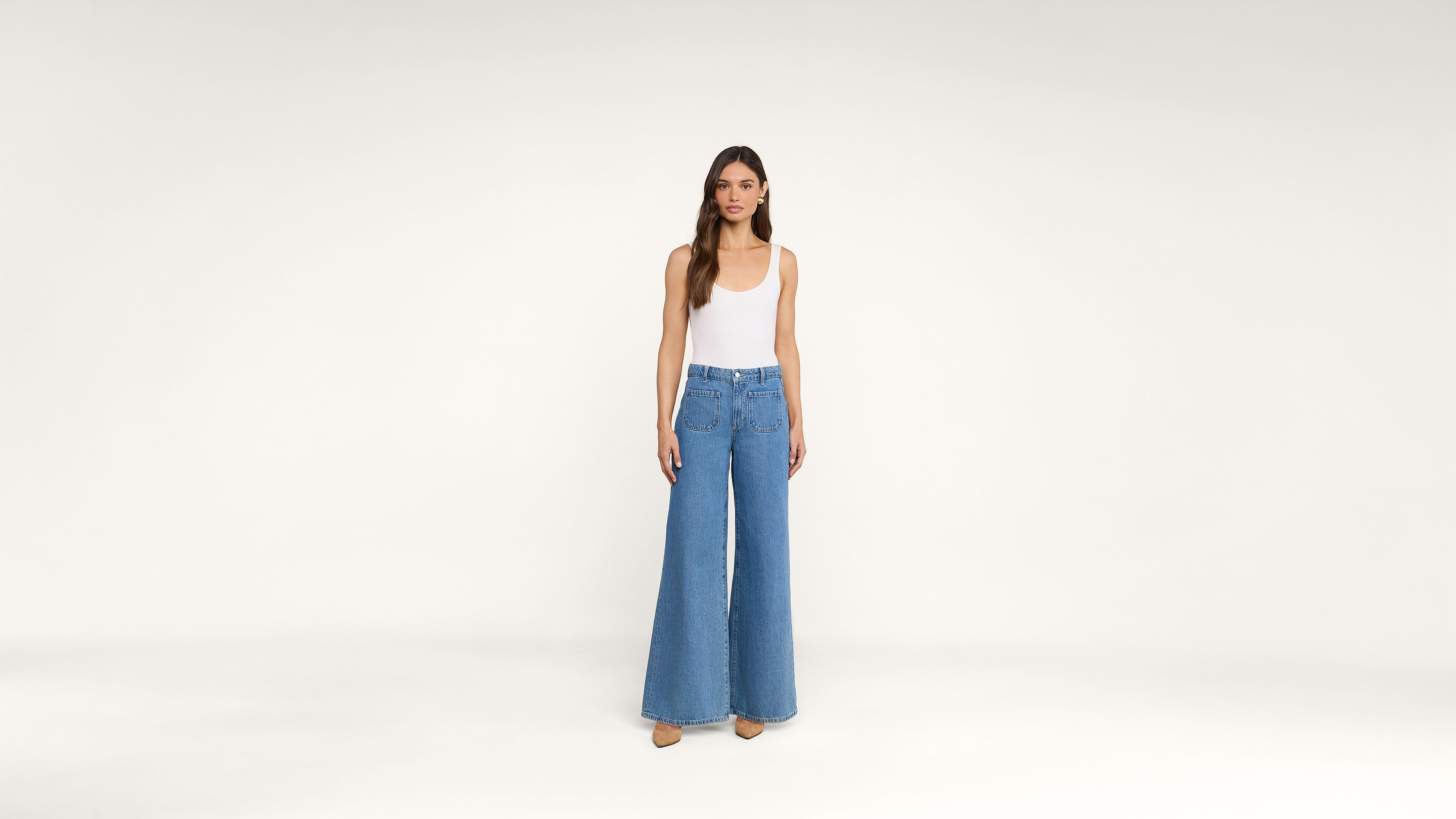 A woman stands against a plain white background, wearing a white sleeveless top and high-waisted, wide-leg blue jeans with beige shoes.