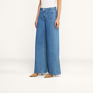 Une personne se tient debout devant un fond beige uni, vêtue dun jean à jambes larges Delcan, dun haut blanc sans manches et descarpins beiges à bout pointu.