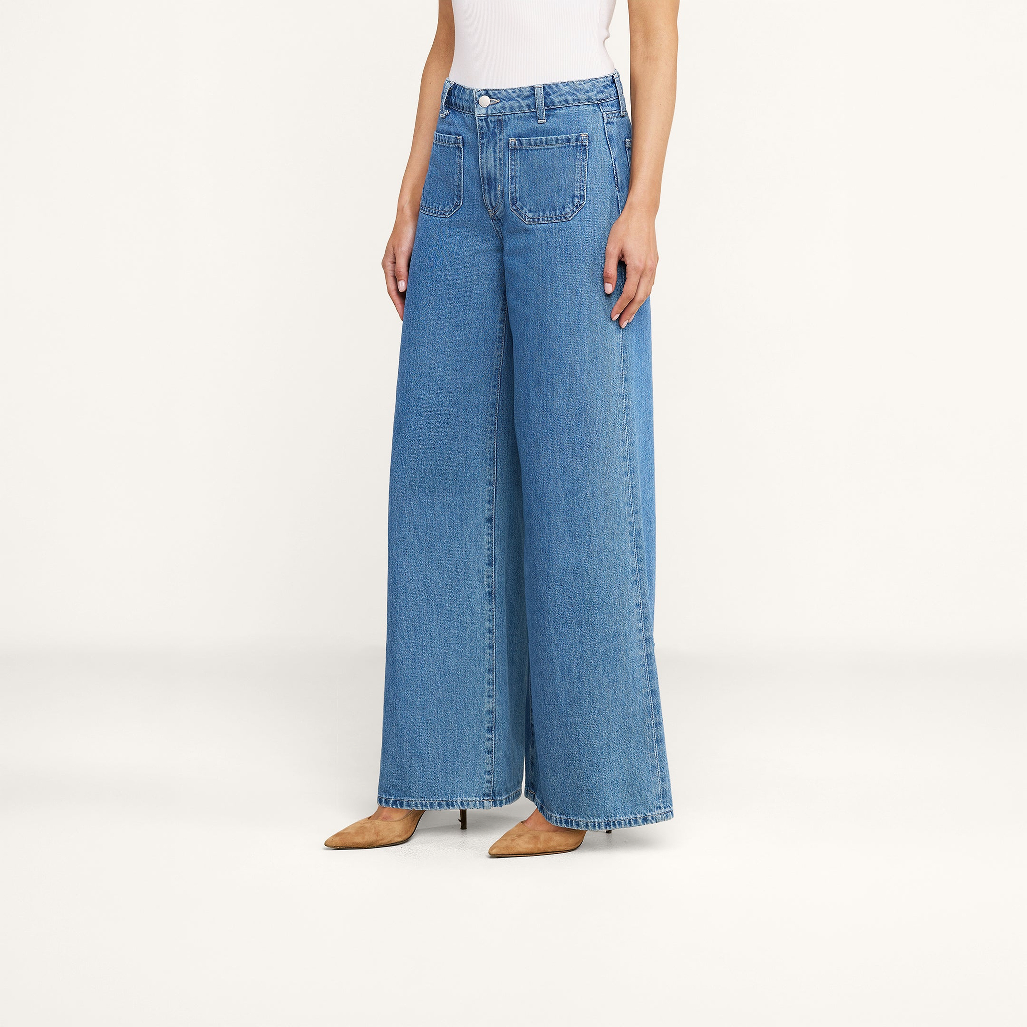 Une personne se tient debout devant un fond beige uni, vêtue dun jean à jambes larges Delcan, dun haut blanc sans manches et descarpins beiges à bout pointu.