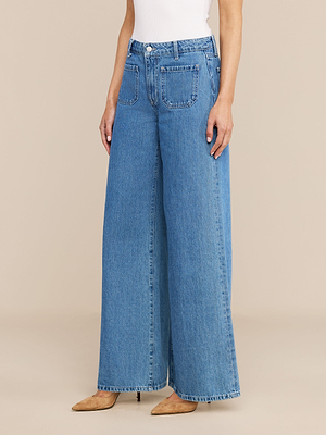 تقف امرأة أمام خلفية بيج سادة، وهي ترتدي بنطال جينز «Delcan Wide-Leg Jean»، وقميصًا أبيض بلا أكمام، وحذاءً بكعب مدبب بلون بني فاتح.