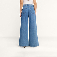 On voit une personne de dos portant le jean à jambes larges Delcan avec des poches plaquées, associé à un haut blanc, sur un fond beige uni. Ce jean taille haute et ample tombe jusquau sol, pour un look décontracté.