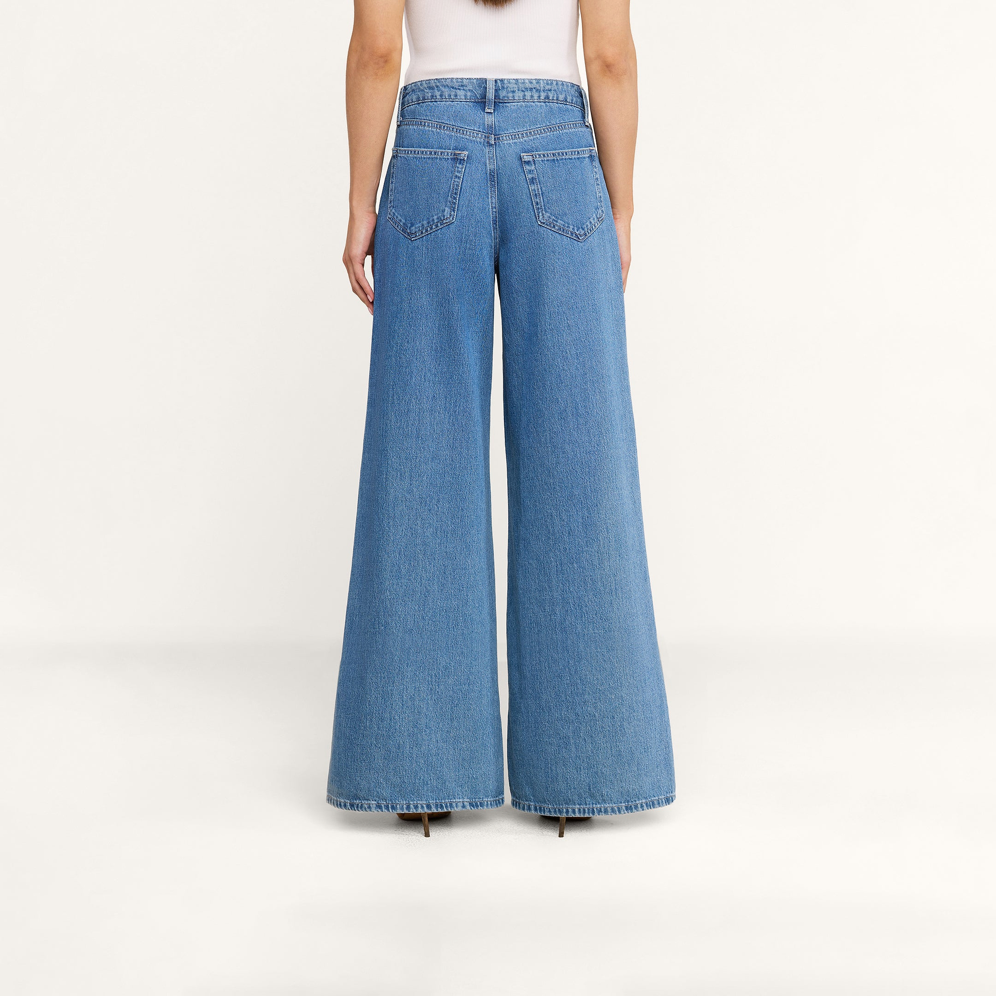 On voit une personne de dos portant le jean à jambes larges Delcan avec des poches plaquées, associé à un haut blanc, sur un fond beige uni. Ce jean taille haute et ample tombe jusquau sol, pour un look décontracté.