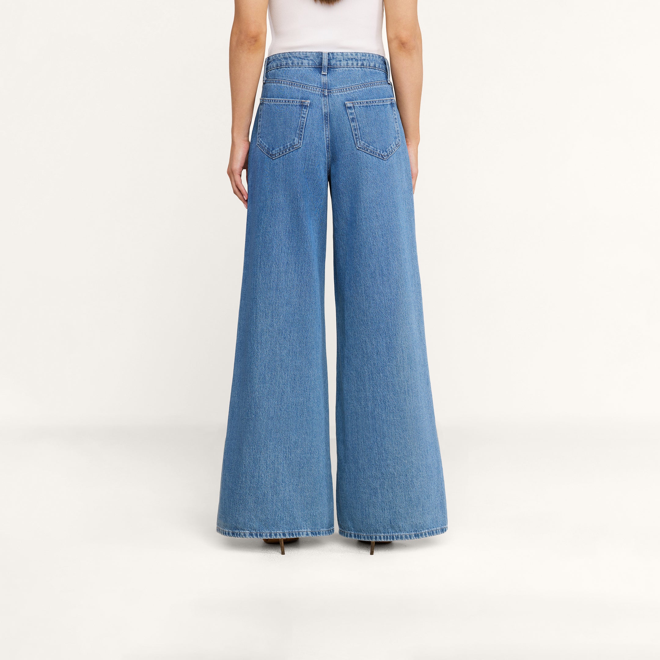 On voit une personne de dos portant le jean à jambes larges Delcan avec des poches plaquées, associé à un haut blanc, sur un fond beige uni. Ce jean taille haute et ample tombe jusquau sol, pour un look décontracté.