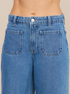 لقطة مقربة لشخص يرتدي بنطال «Delcan Wide-Leg Jean»، وهو بنطال من الدنيم الأزرق ذو خصر مرتفع، ويحتوي على جيبين أماميين ملتصقين، وإغلاق بأزرار، وحلقات للحزام؛ ويظهر بطنه المكشوف فوق حزام البنطال.