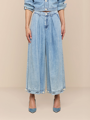 Une personne porte le jean à plis Elias, une veste en jean assortie et des talons bleus à bouts pointus, sur un fond beige. Limage est coupée au niveau de la poitrine.