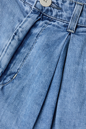 Gros plan sur le jean à jambes larges Elias en denim poids plume bleu clair, avec des détails plissés sur le devant, des coutures apparentes et un bouton argenté à la taille - une pièce de transition idéale.