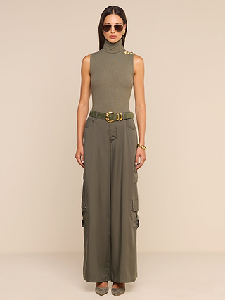 A woman stands facing forward, wearing sunglasses, a sleeveless olive turtleneck top with buttons on the shoulder, and the Isolde Cargo Pant—lightweight Tencel wide-leg cargo pants cinched with a belt. She has long hair and is standing against a plain beige background.