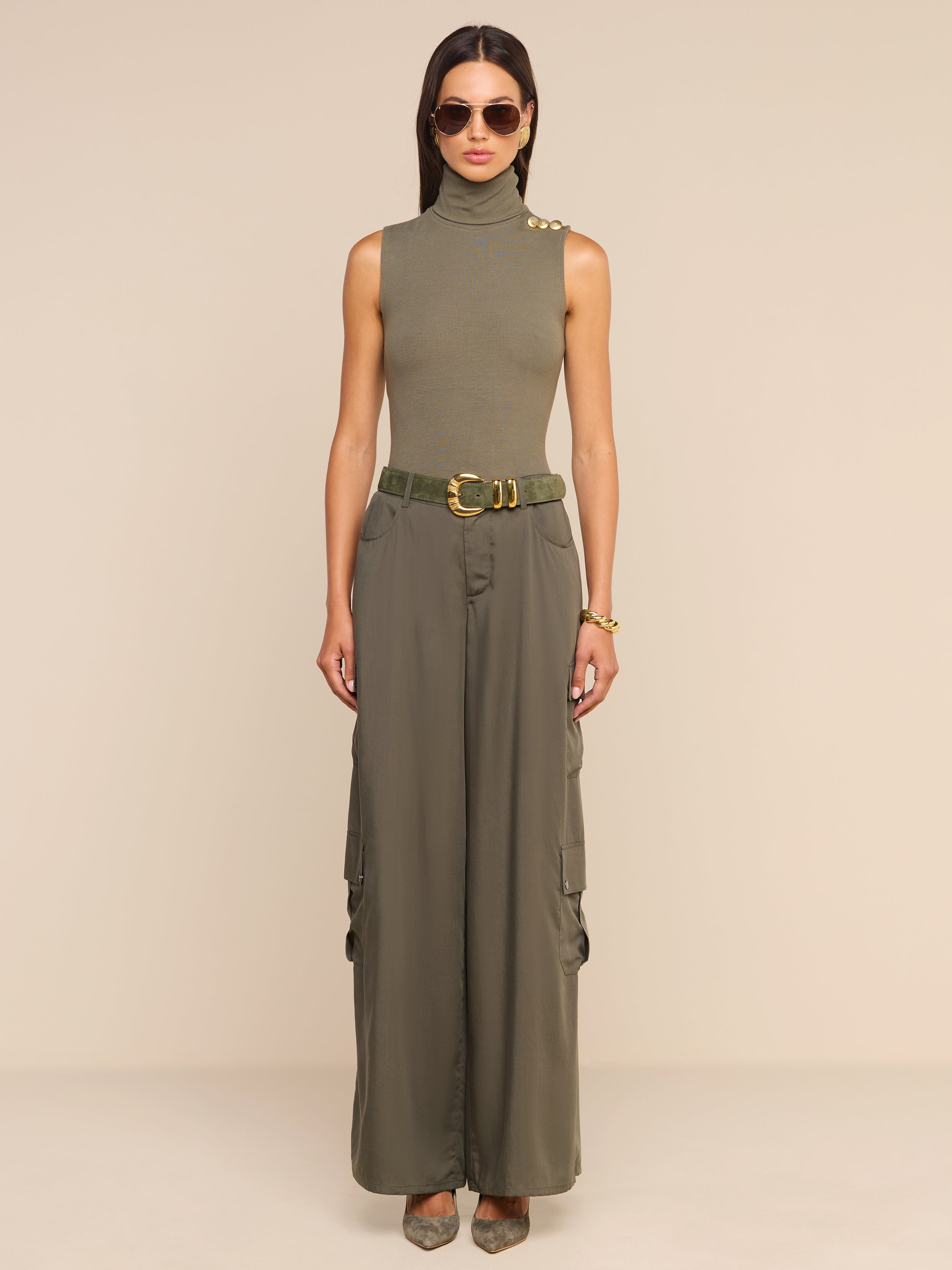 A woman wearing sunglasses, a sleeveless olive green turtleneck top, the Isolde Cargo Pant crafted from lightweight Tencel, a belt, and heels stands against a plain white background.