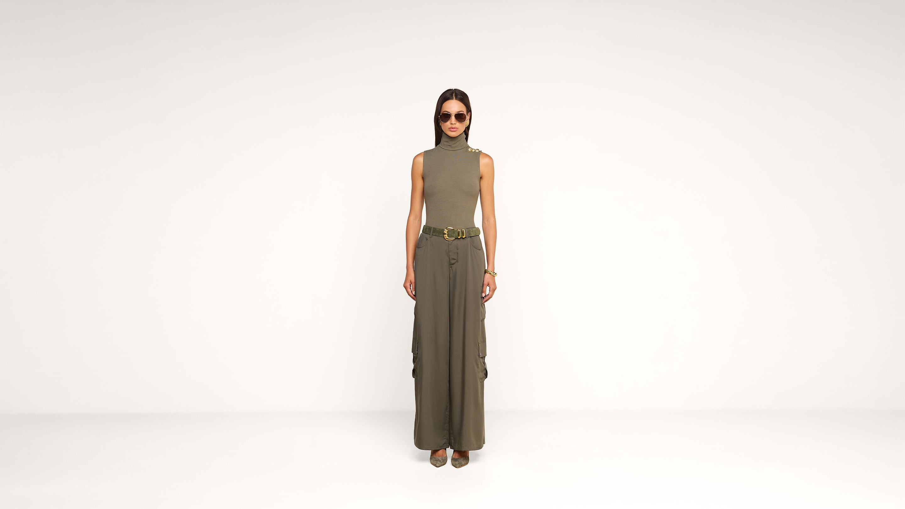 A woman stands against a plain white background, wearing sunglasses, a sleeveless turtleneck top, wide-leg cargo pants, a belt, and heeled shoes. She has straight hair and her hands are by her sides.