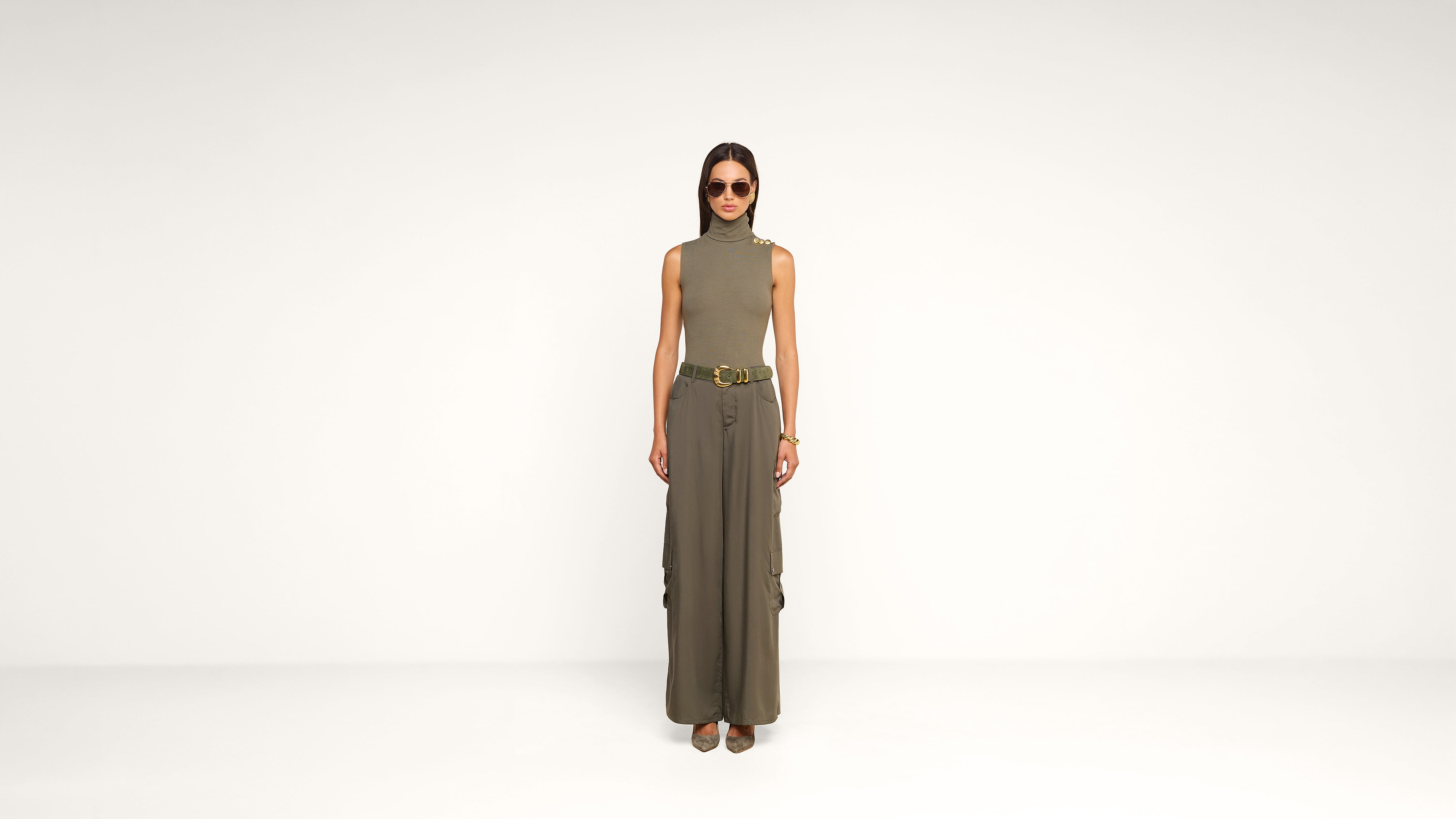 A woman stands against a plain white background, wearing sunglasses, a sleeveless turtleneck top, wide-leg cargo pants, a belt, and heeled shoes. She has straight hair and her hands are by her sides.