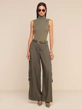A woman wearing sunglasses, a sleeveless olive turtleneck, and the Isolde Cargo Pant with heels stands against a beige background. She accessorizes with a belt and bracelets.