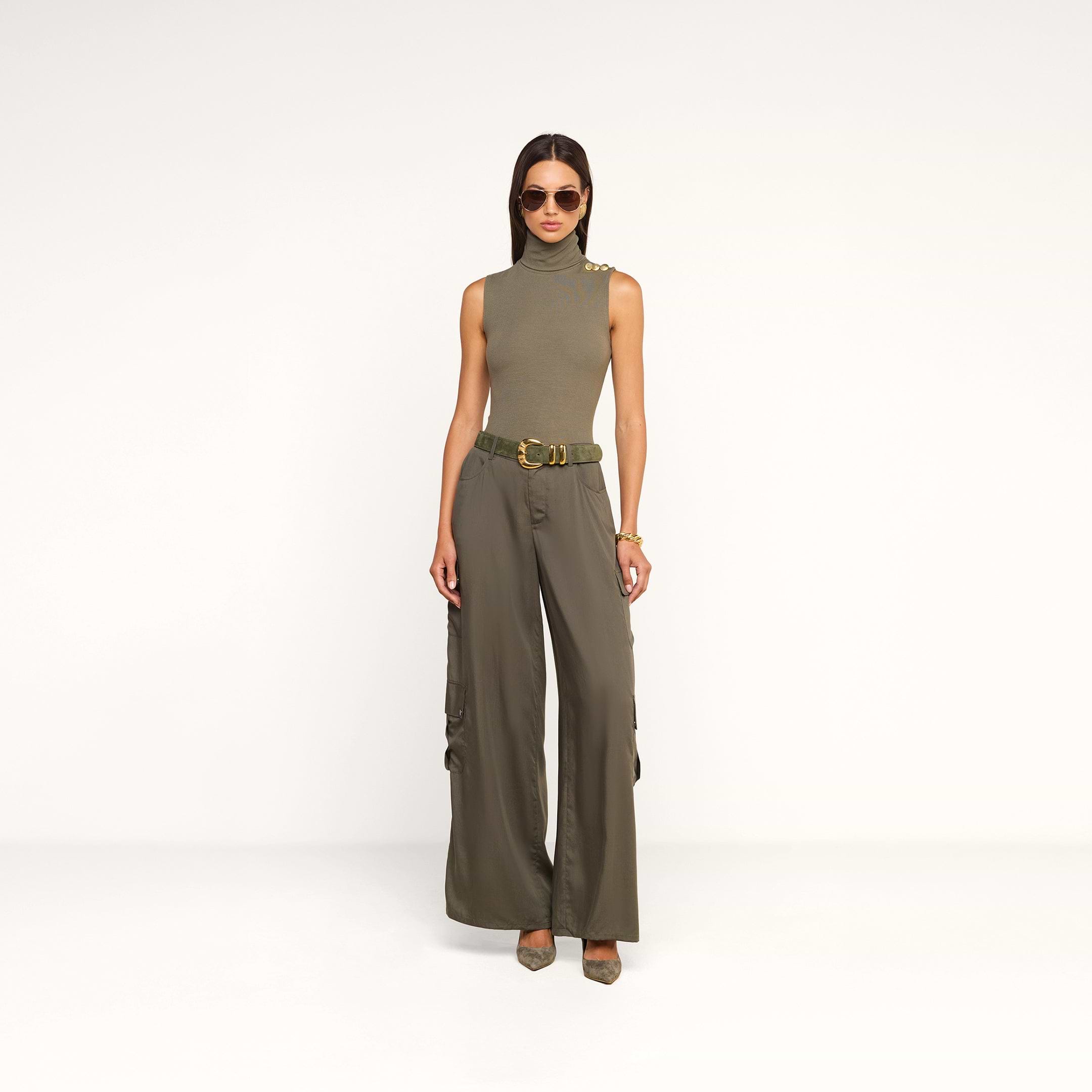 A woman wearing sunglasses, a sleeveless olive-green turtleneck top, and the Isolde Cargo Pant with pointed shoes stands against a plain white background.