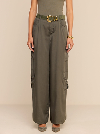 A person wearing the Isolde Cargo Pant—high-waisted, wide-leg olive cargo pants crafted from lightweight tencel with side pockets—paired with a green belt featuring a gold buckle, a gold bracelet, and pointed shoes stands against a plain beige background.