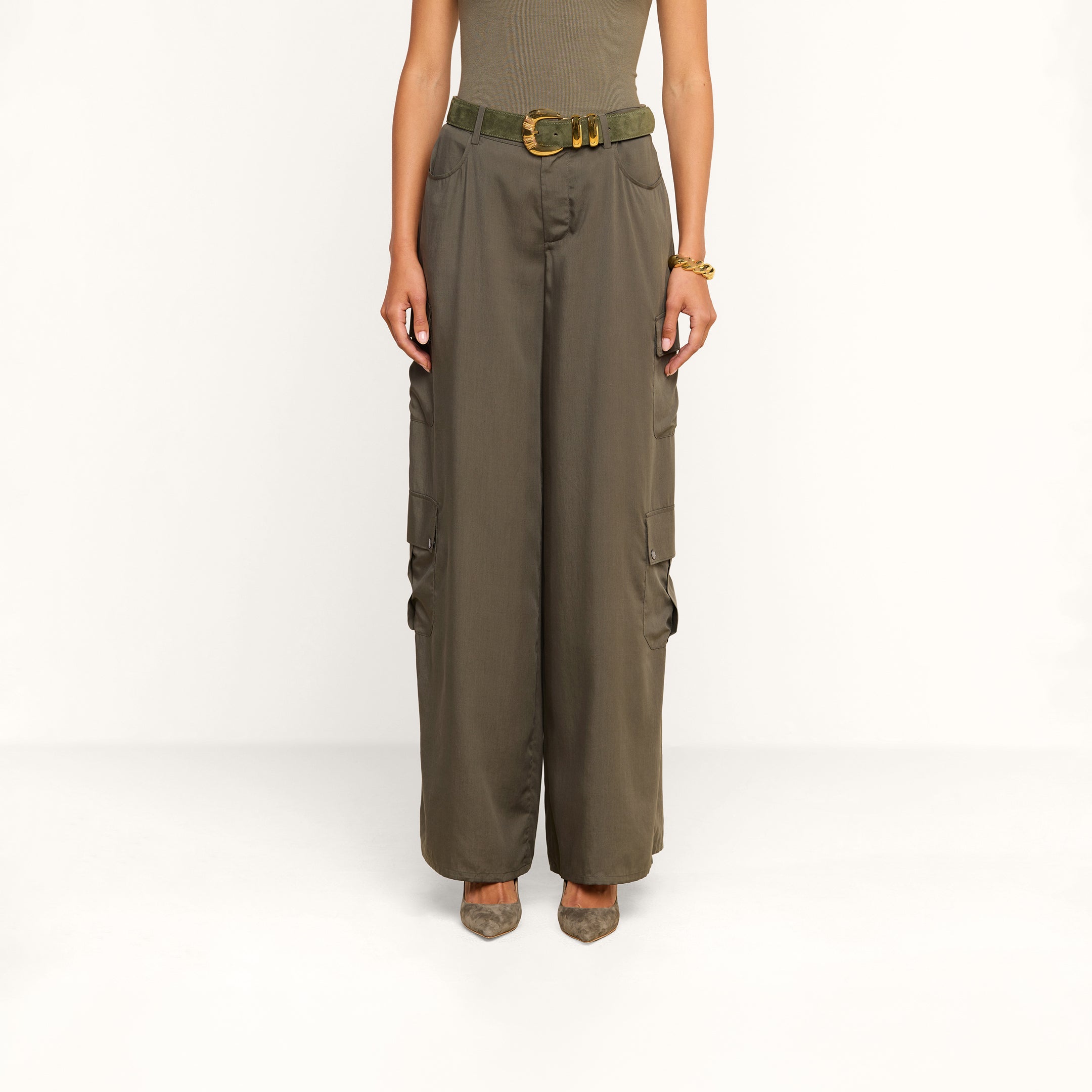 Wearing the Isolde Cargo Pant—high-waisted, wide-leg olive cargo pants made from lightweight Tencel and finished with a matching belt—a person pairs them with a sleeveless top and pointed shoes while standing against a plain white background.