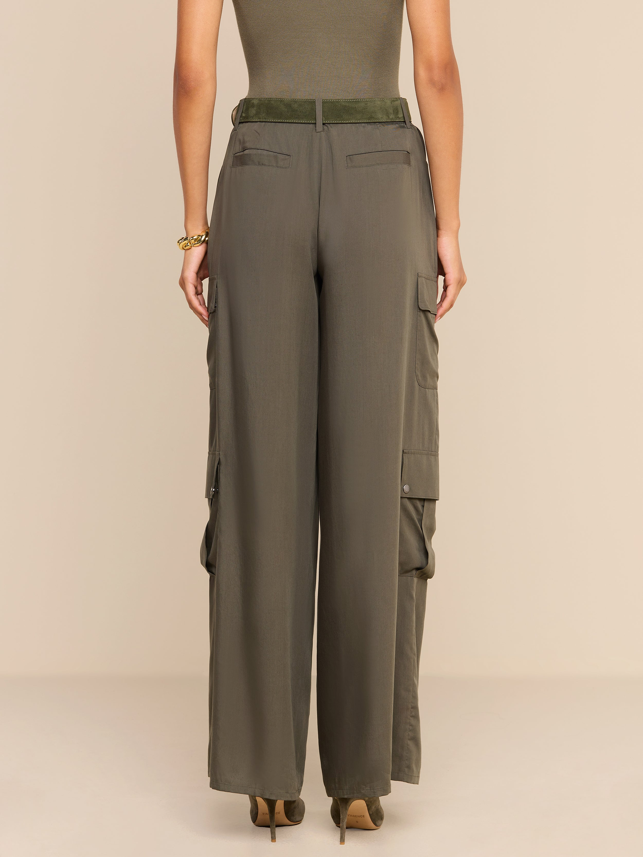 A woman stands facing away, wearing the Isolde Cargo Pant—high-waisted, olive-green wide-leg cargo pants crafted from lightweight tencel. She pairs them with a sleeveless top and heeled shoes. Her arms rest relaxed by her sides against a plain, light background.