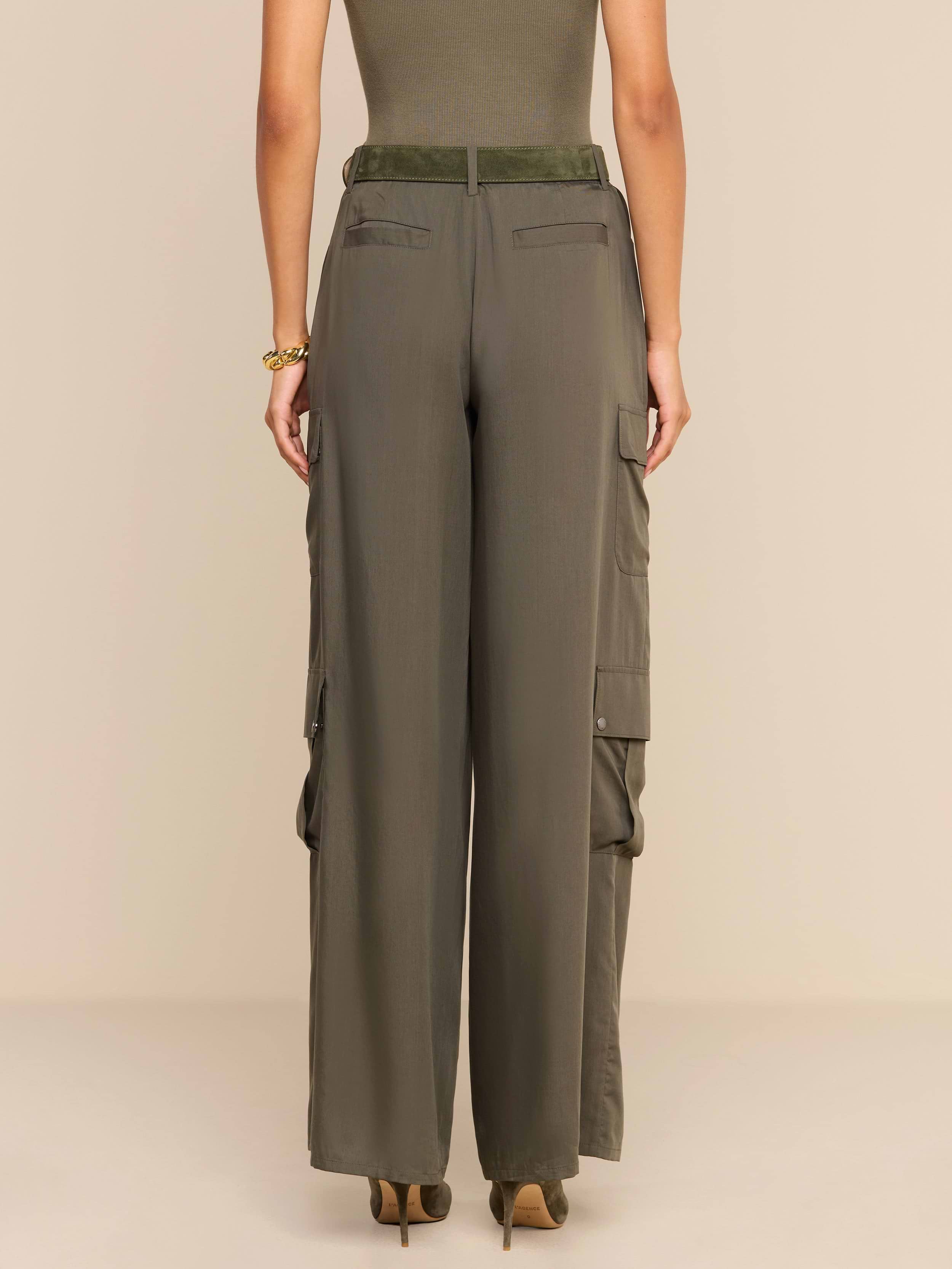 A woman stands facing away, wearing the Isolde Cargo Pant—high-waisted, olive-green wide-leg cargo pants crafted from lightweight tencel. She pairs them with a sleeveless top and heeled shoes. Her arms rest relaxed by her sides against a plain, light background.