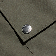 Close-up of an olive green fabric pocket with a metal snap button, featuring small engraved text around its edge. The lightweight Tencel texture and visible stitching highlight the Isolde Cargo Pant’s design.