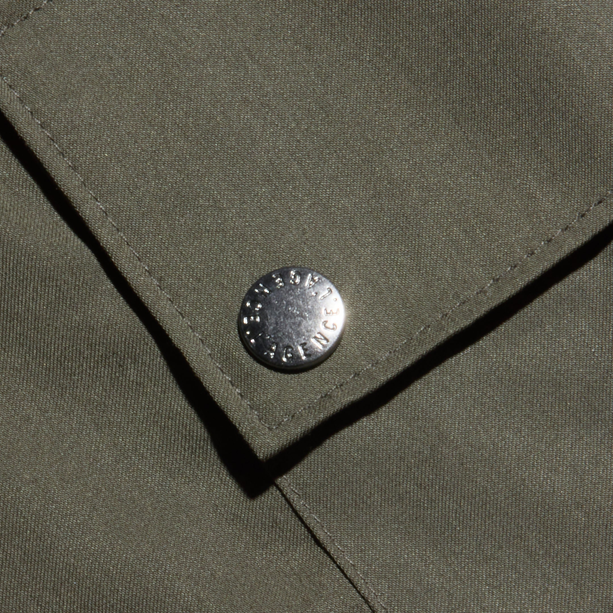 Close-up of an olive green fabric pocket with a metal snap button, featuring small engraved text around its edge. The lightweight Tencel texture and visible stitching highlight the Isolde Cargo Pant’s design.