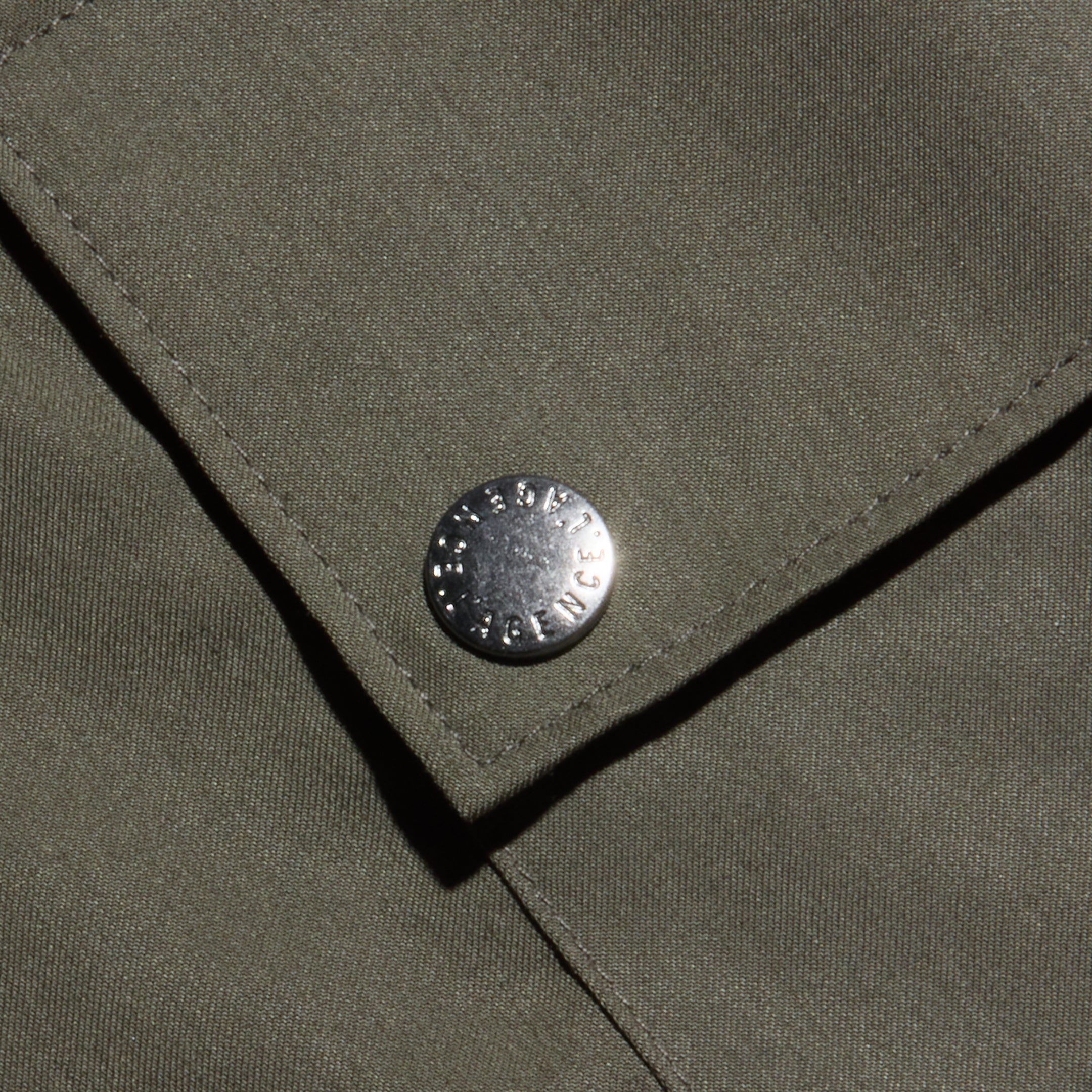 Close-up of an olive green fabric pocket with a metal snap button, featuring small engraved text around its edge. The lightweight Tencel texture and visible stitching highlight the Isolde Cargo Pant’s design.