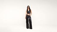 A woman in black sunglasses, a sleeveless black leather crop top, and black wide-leg leather pants stands against a plain white background.