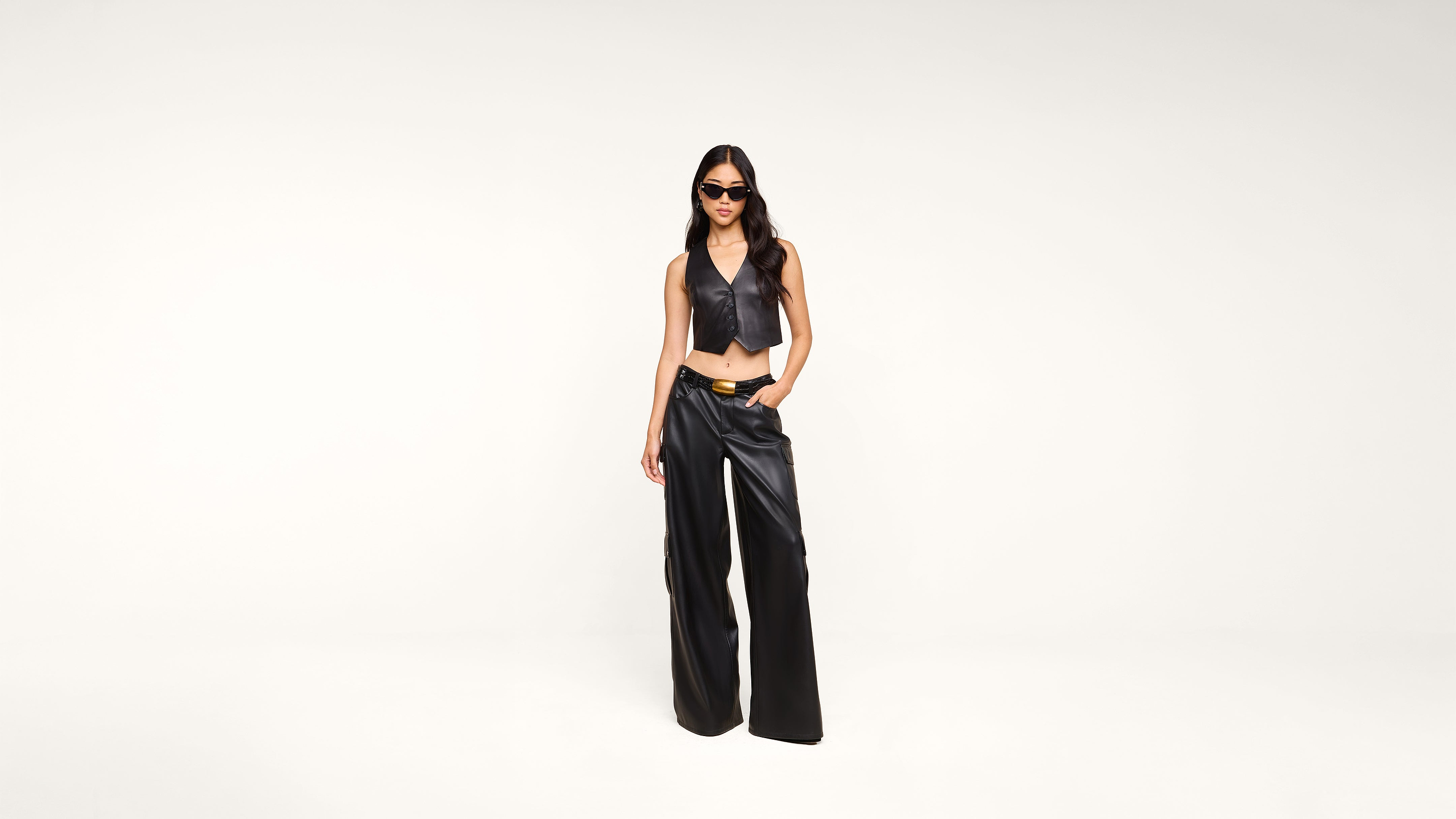 A woman in black sunglasses, a sleeveless black leather crop top, and black wide-leg leather pants stands against a plain white background.