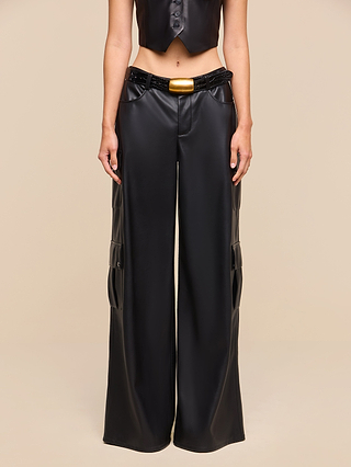 A person wearing the Isolde Cargo Pant—a pair of wide-leg vegan leather cargo pants with utility pockets—styled with a black cropped vest and a black belt featuring a large gold buckle, stands against a beige background.