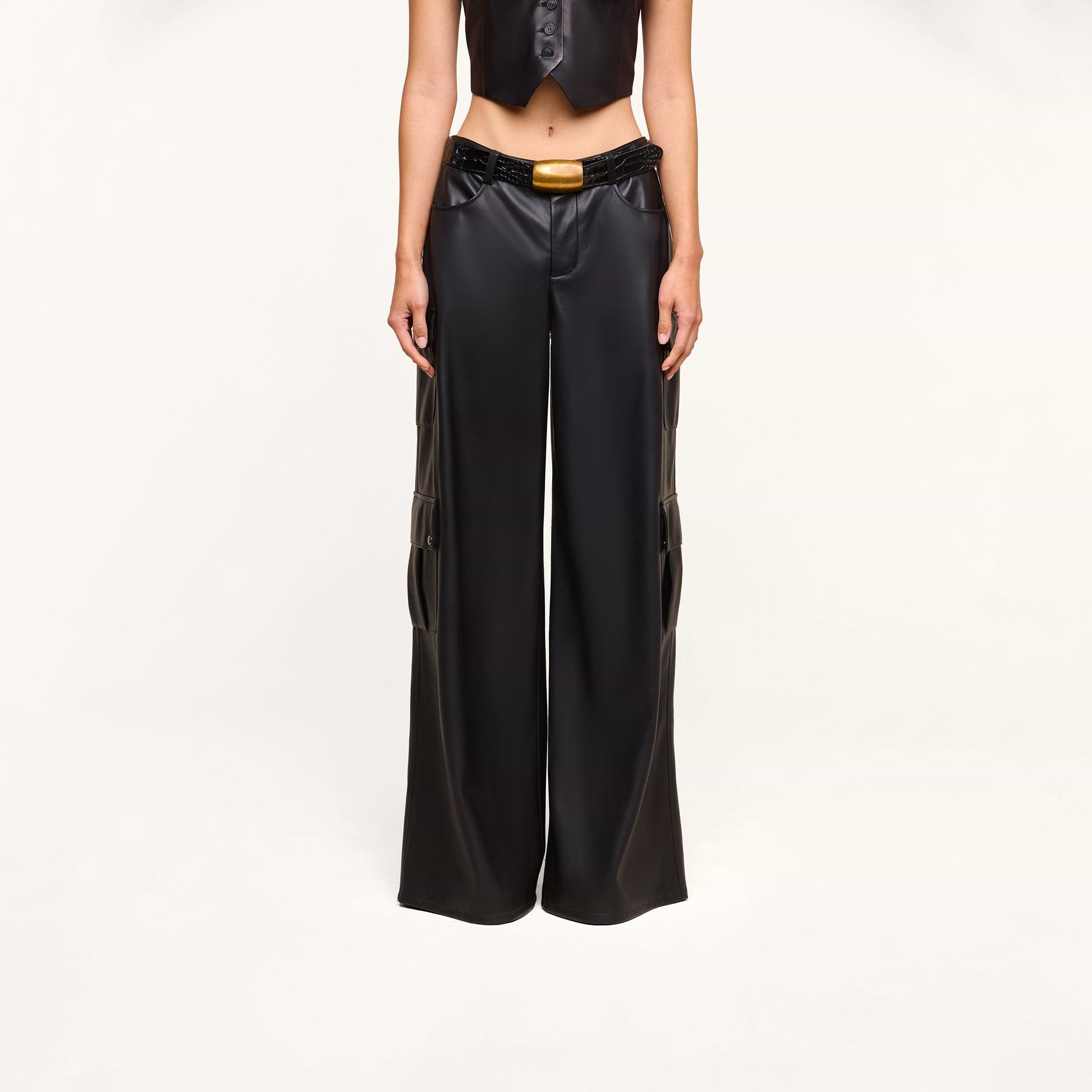 Wearing the Isolde Cargo Pant—black wide-leg vegan leather pants with utility pockets and a belt featuring a yellow buckle—paired with a matching cropped black vest, this person stands against a plain white background with their face out of frame.