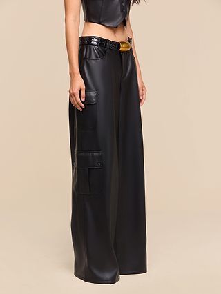 Wearing the Isolde Cargo Pant—high-waisted, wide-leg vegan leather cargo pants with utility pockets and a black belt—paired with a black sleeveless leather top, a person stands against a beige background.