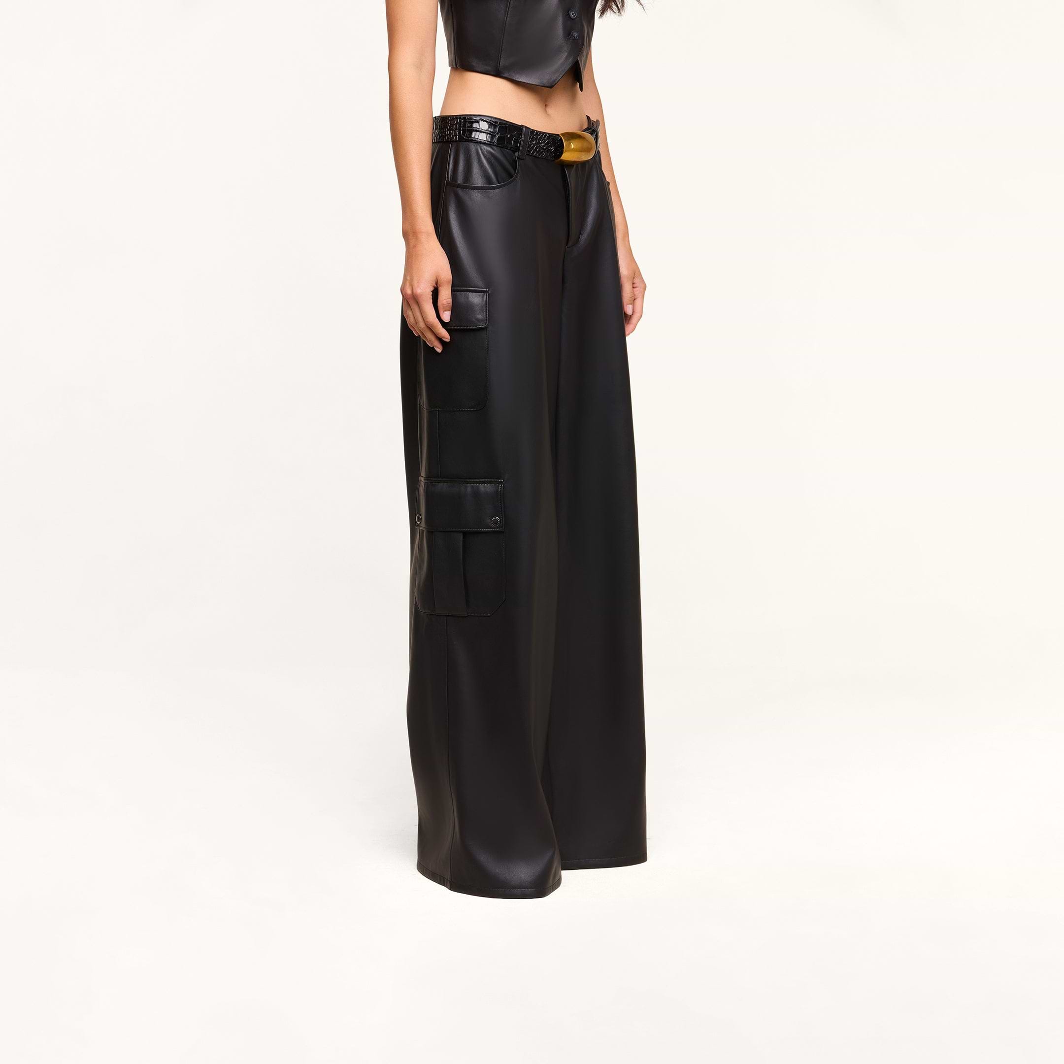 A person wearing the Isolde Cargo Pant—high-waisted black wide-leg vegan leather cargo pants with utility pockets—paired with a black crop top, standing against a plain white background.