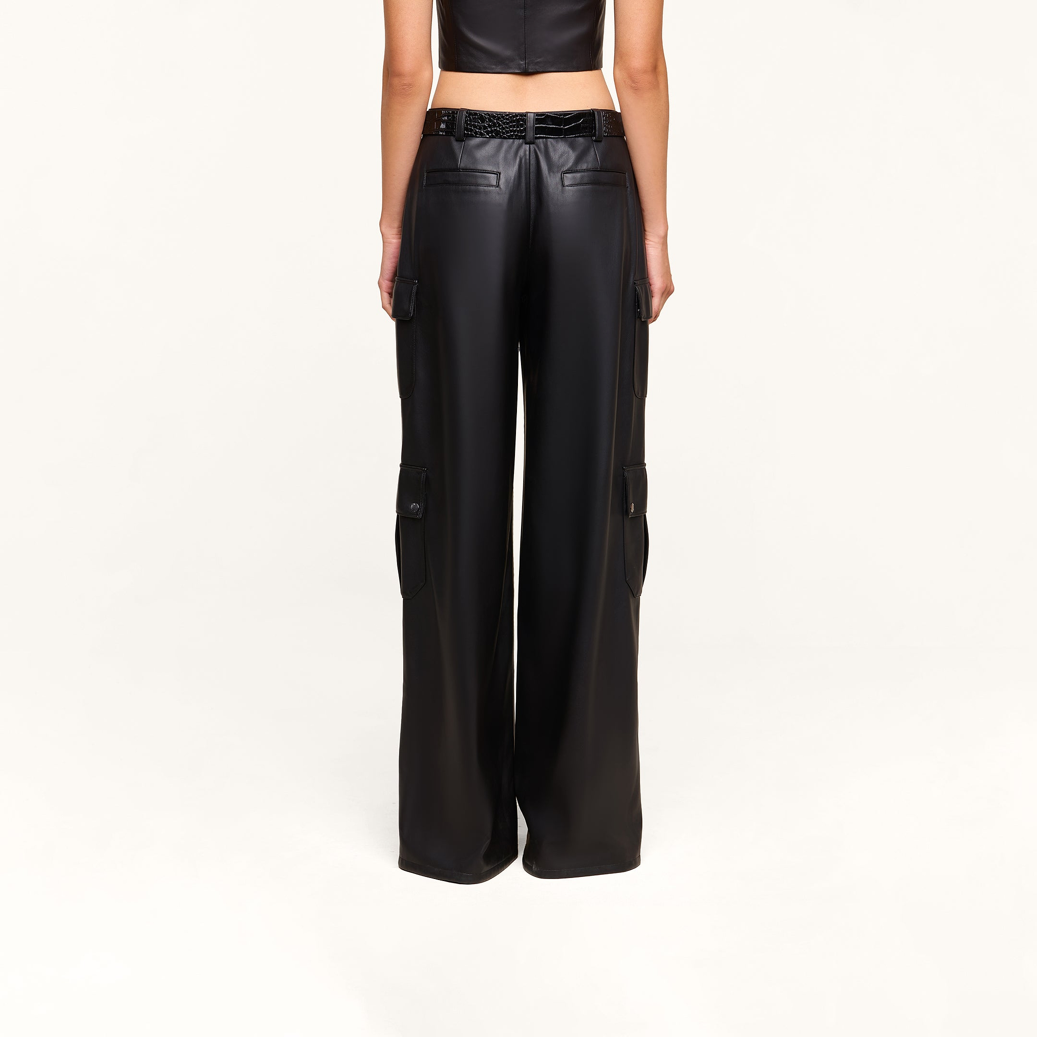 A person is shown from the back against a plain light background, wearing the Isolde Cargo Pant—a high-waisted, black, wide-leg vegan leather cargo pant with utility pockets—paired with a cropped sleeveless black top.