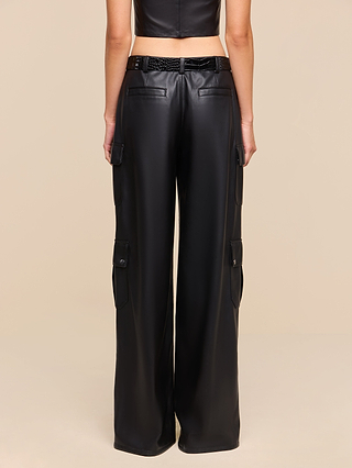 Wearing the Isolde Cargo Pant—a high-waisted black vegan leather cargo featuring utility pockets and a black belt—the person is shown from the back against a plain beige background.