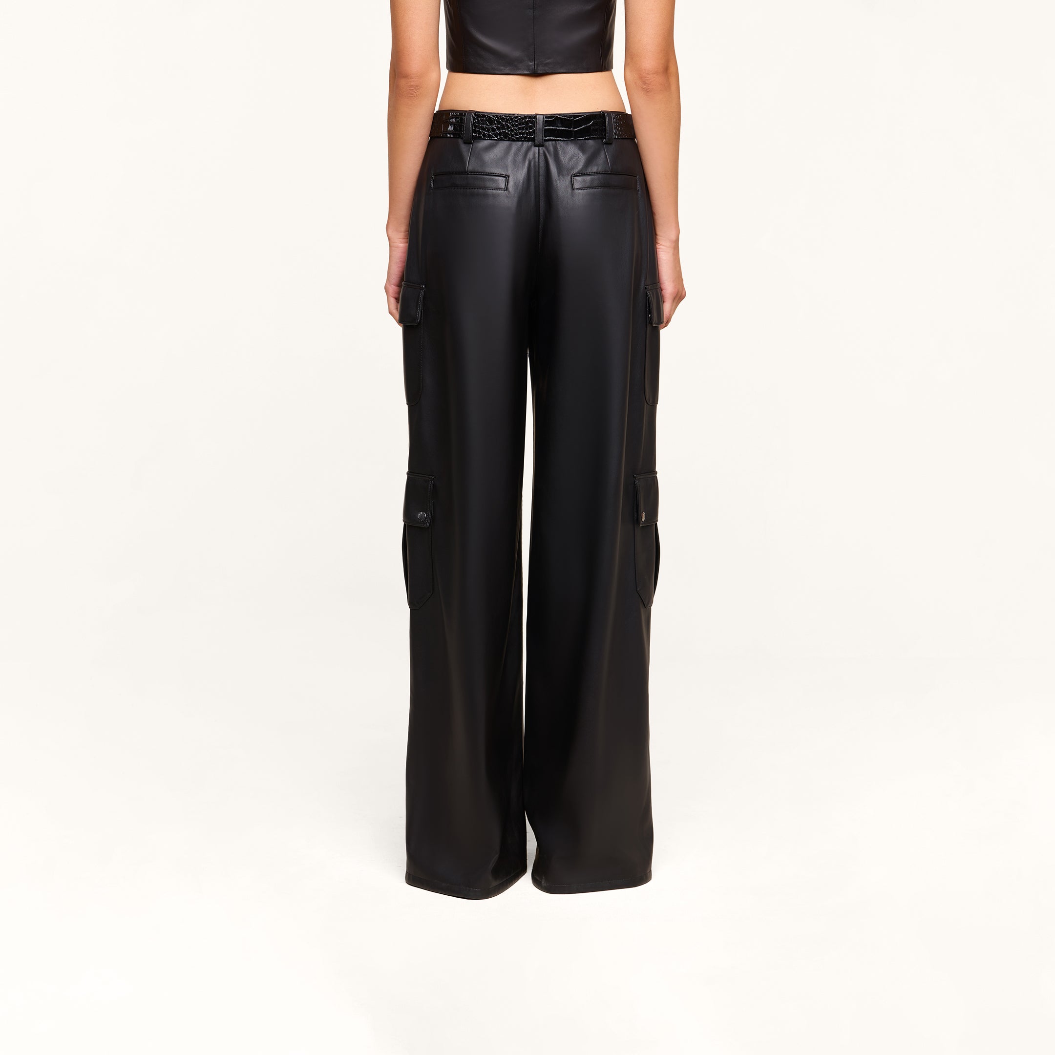 A person is shown from the back against a plain light background, wearing the Isolde Cargo Pant—a high-waisted, black, wide-leg vegan leather cargo pant with utility pockets—paired with a cropped sleeveless black top.