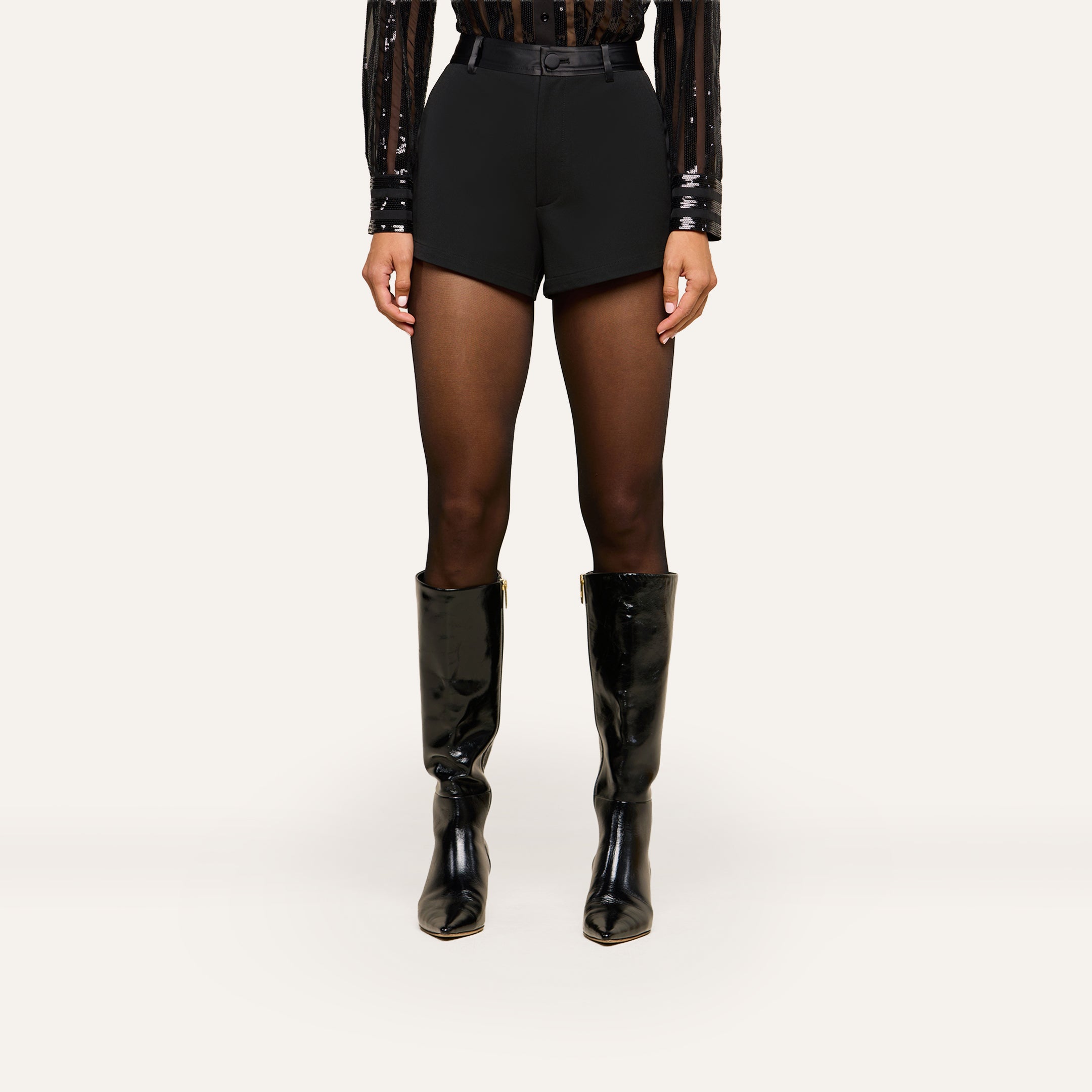 Wearing a black sequin long-sleeve top, sheer black tights, and shiny black knee-high boots, the person showcases the Colt Tuxedo Short—black high-waisted shorts crafted from suiting fabric—while standing against a plain light background.